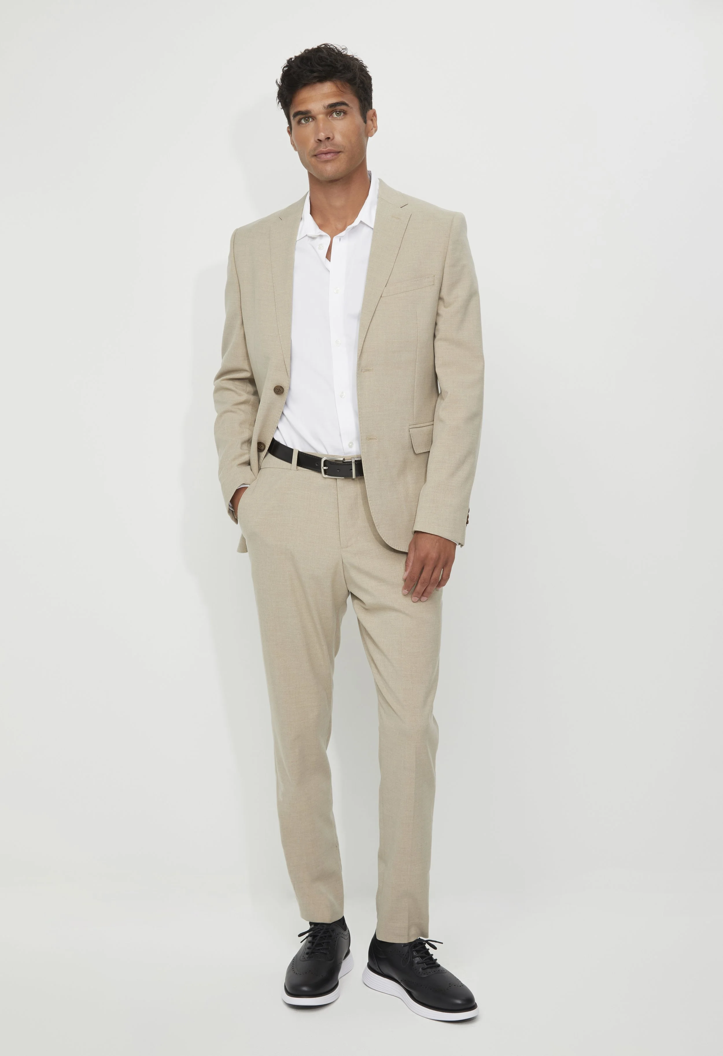 Man in beige suit with white shirt and black sneakers, standing against a plain background.