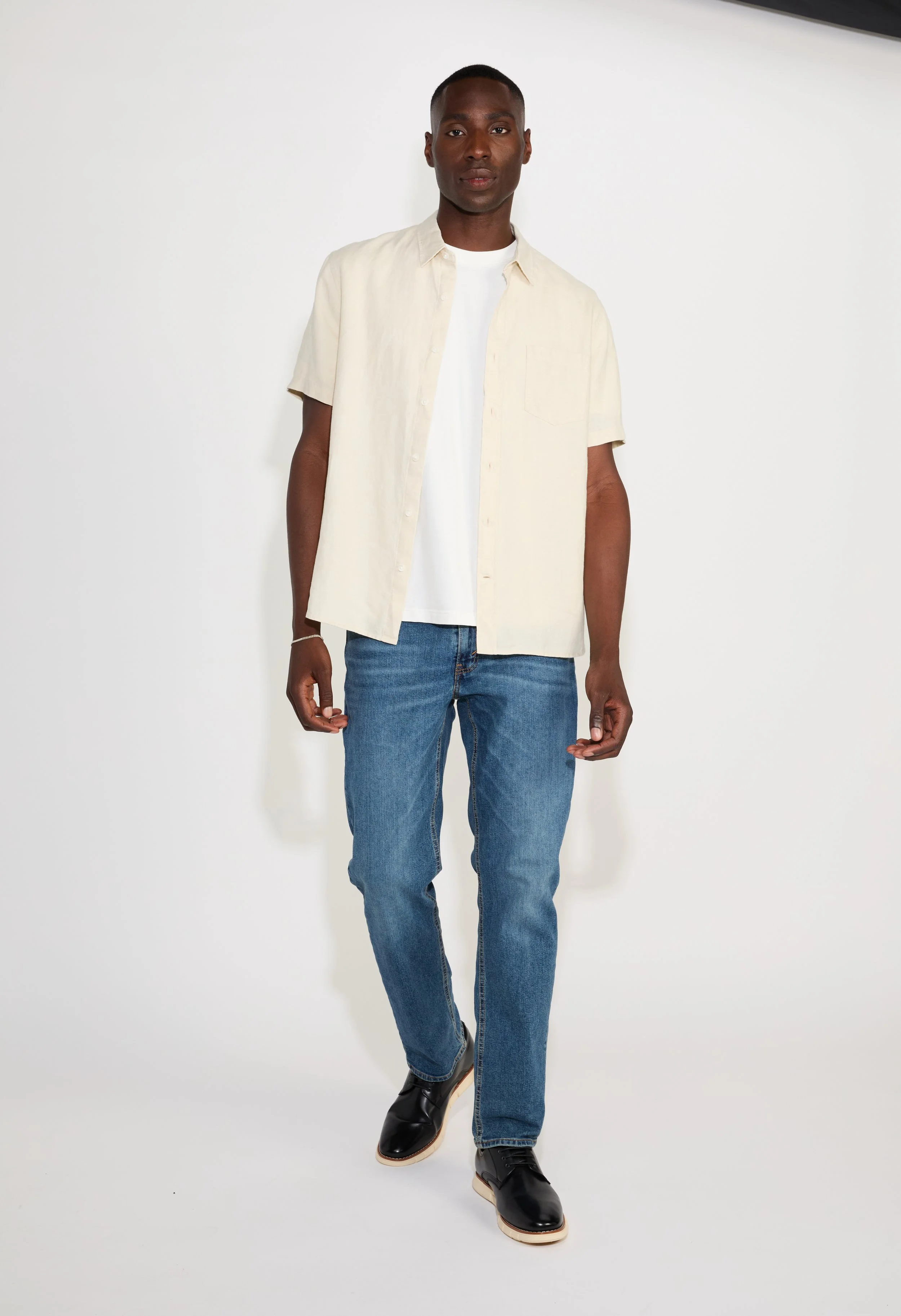 A young man with dark skin and short hair standing against a plain white background, wearing a cream short-sleeved button-up shirt over a white t-shirt, blue jeans, and black shoes.