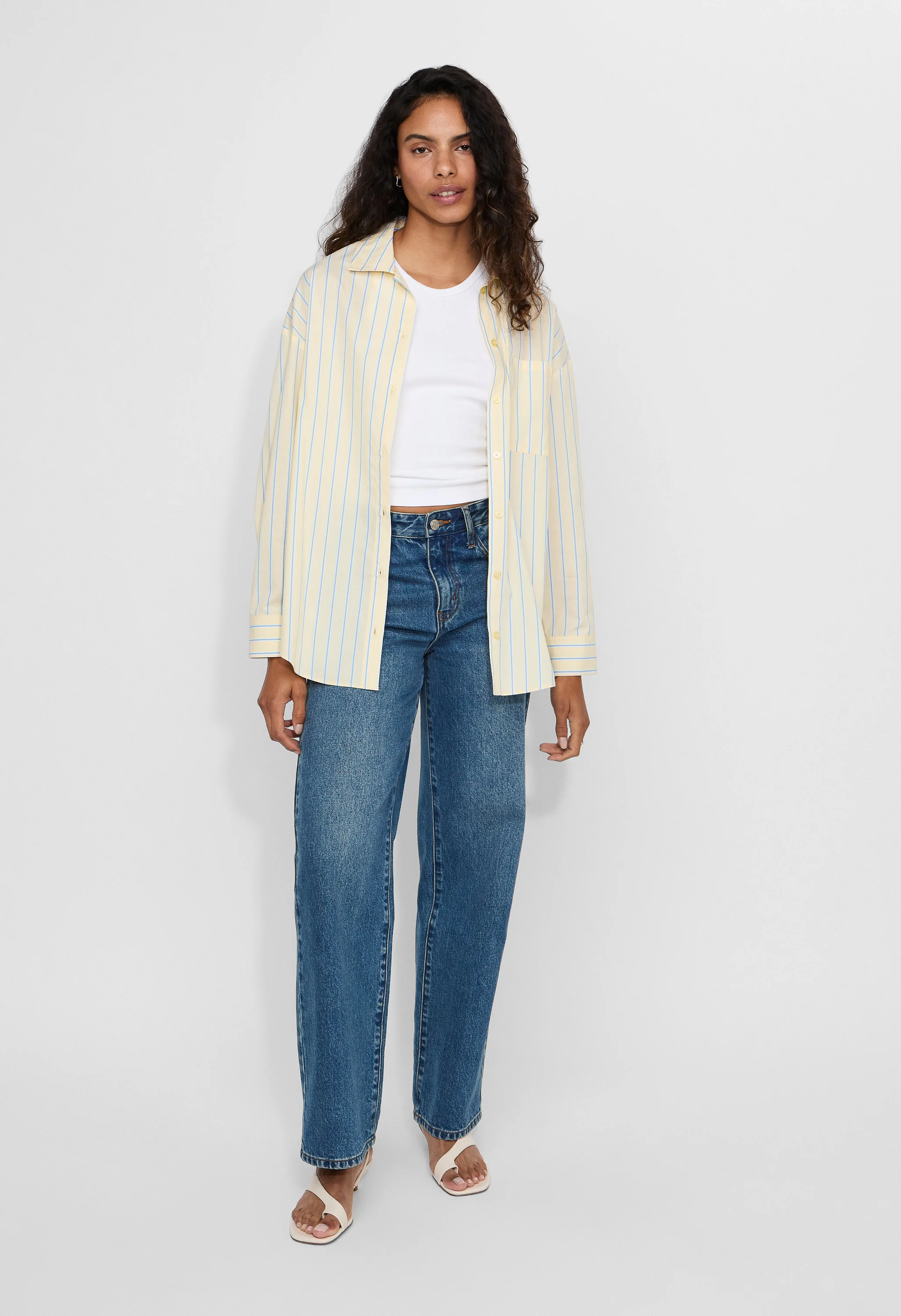 Woman wearing a yellow and blue striped shirt over a white T-shirt, blue jeans, and open-toe heels, standing against a plain white background.