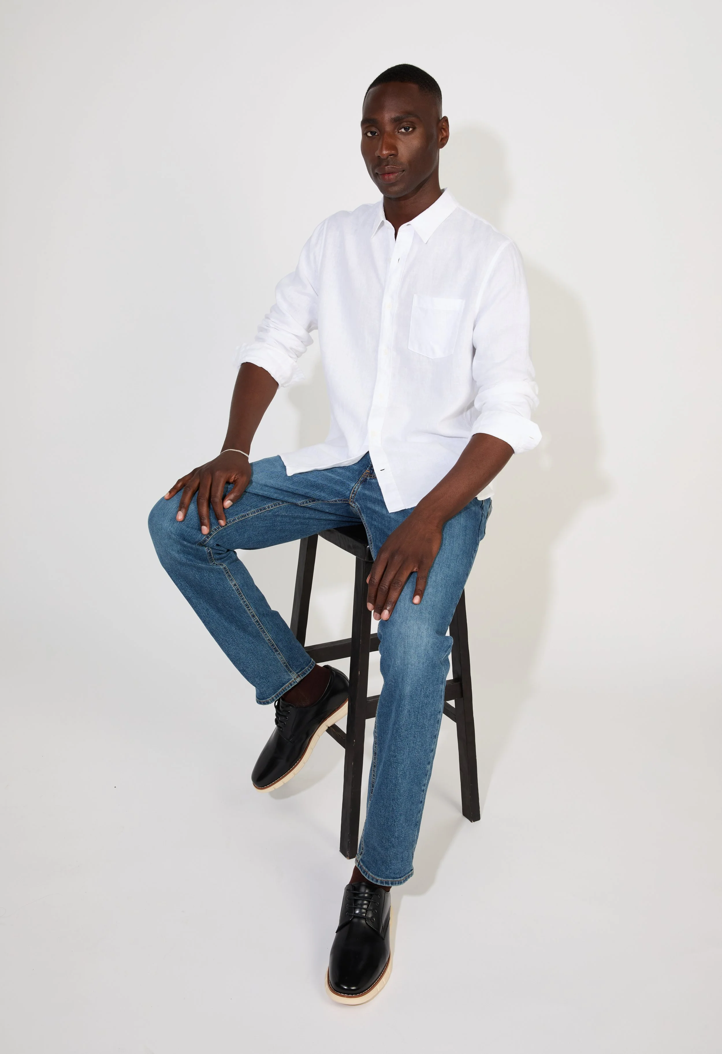 A young Black man with short hair dressed in a white button-up shirt and blue jeans, seated on a black wooden stool against a plain white background.