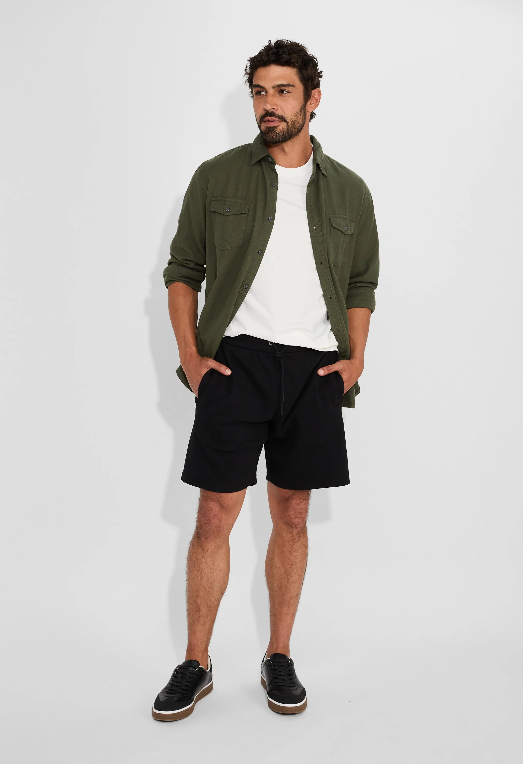 A man with a beard and curly hair wearing a dark green button-up shirt over a white T-shirt, black shorts, and black sneakers, standing against a plain white wall with his hands in his pockets.