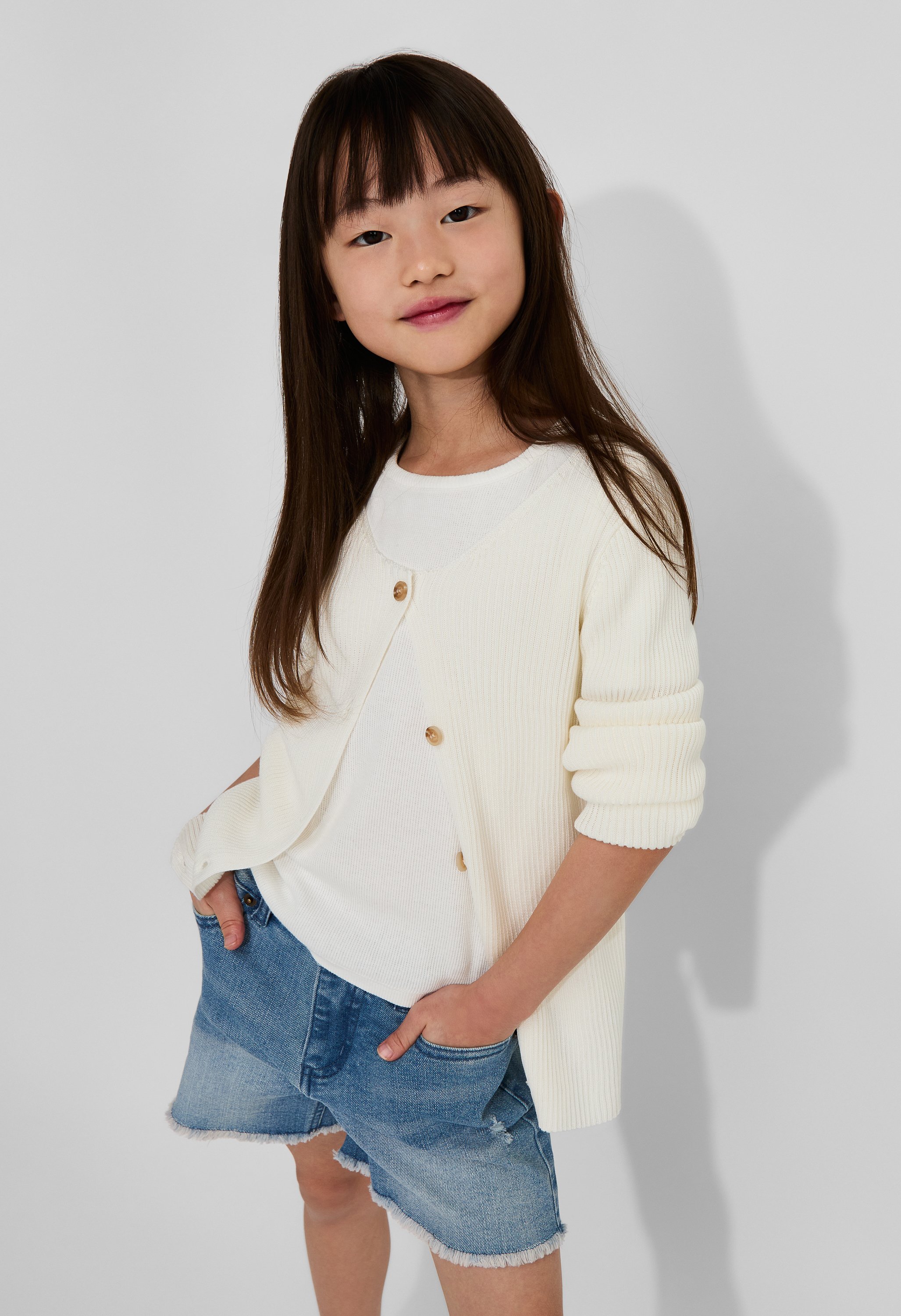Young girl with long brown hair, wearing a cream cardigan over a white top and denim shorts, standing against a plain white background.