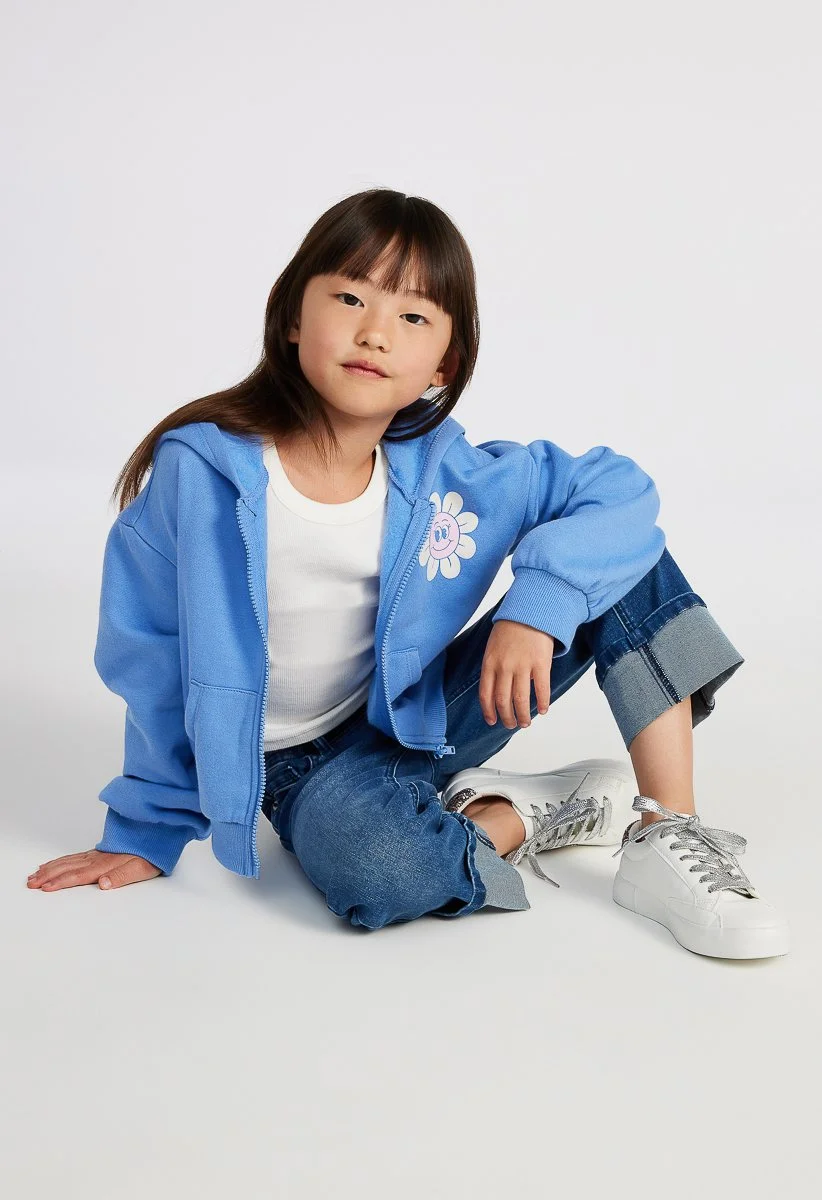 A young girl with long brown hair sitting on the floor against a white background, wearing a blue zip-up hoodie with a smiling flower on it, white t-shirt, rolled-up jeans, and white sneakers.