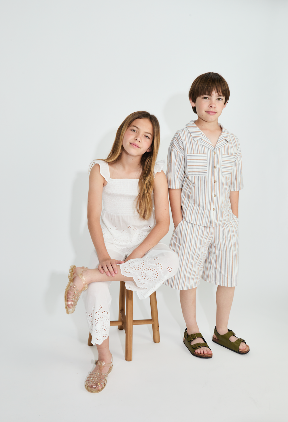A young girl sitting on a stool and a young boy standing next to her against a plain white background.
