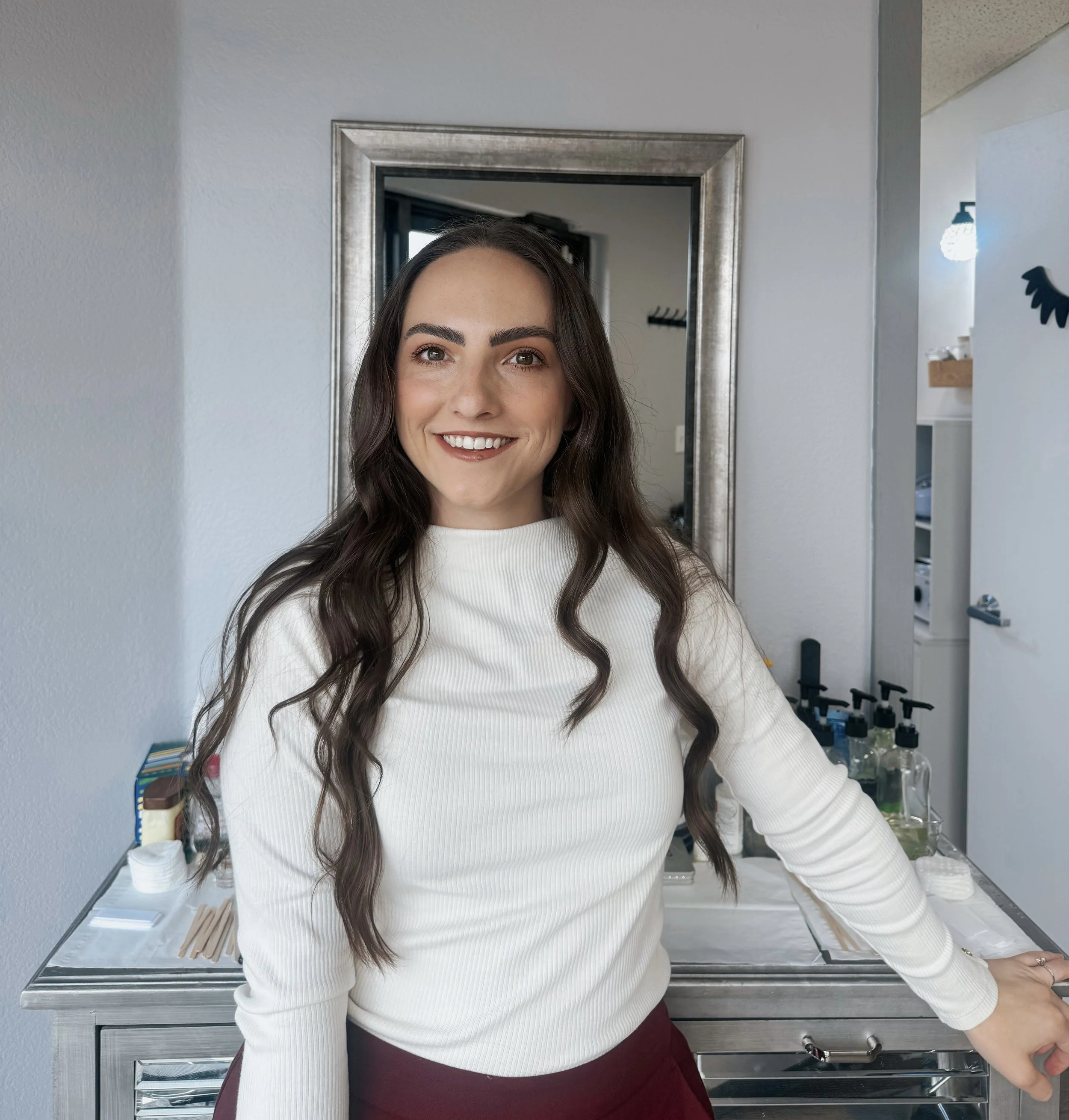 A young woman with long brown hair, wearing a white top, smiling at the camera. She is standing in a kitchen with a stainless steel countertop and shelves with various bottles and kitchen items. There is a silver-framed mirror behind her reflecting part of the kitchen.