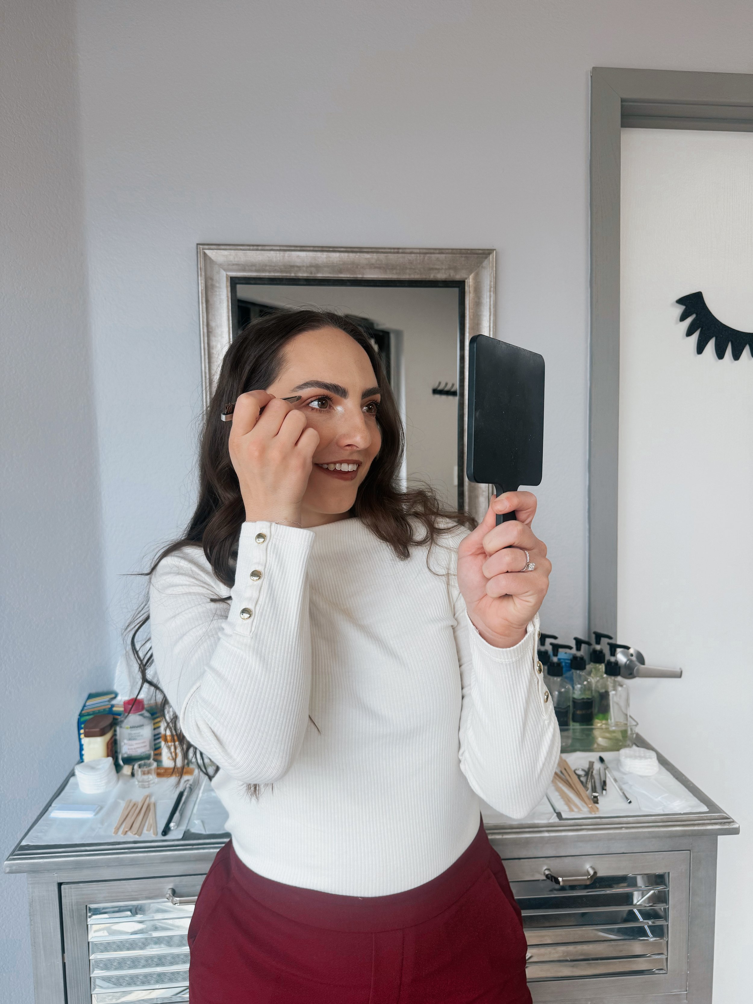 Woman with brown hair and a white sweater applying makeup in front of a mirror, holding a mirror in her left hand, standing in a room with a metal medical or beauty workstation in the background.