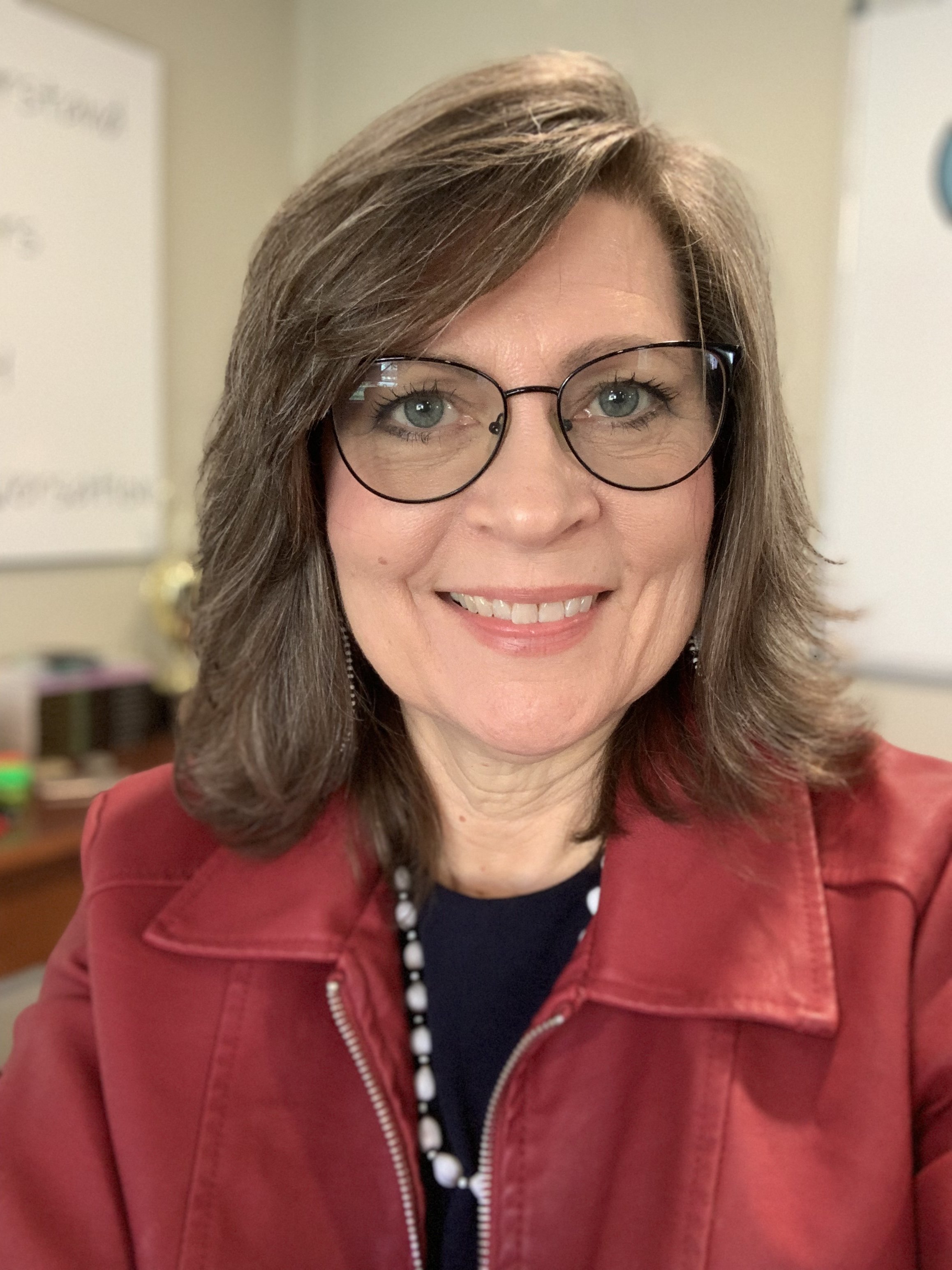 Close-up of a smiling woman with light brown hair, wearing glasses, a red jacket, and a black top with white polka dots, in an indoor setting.