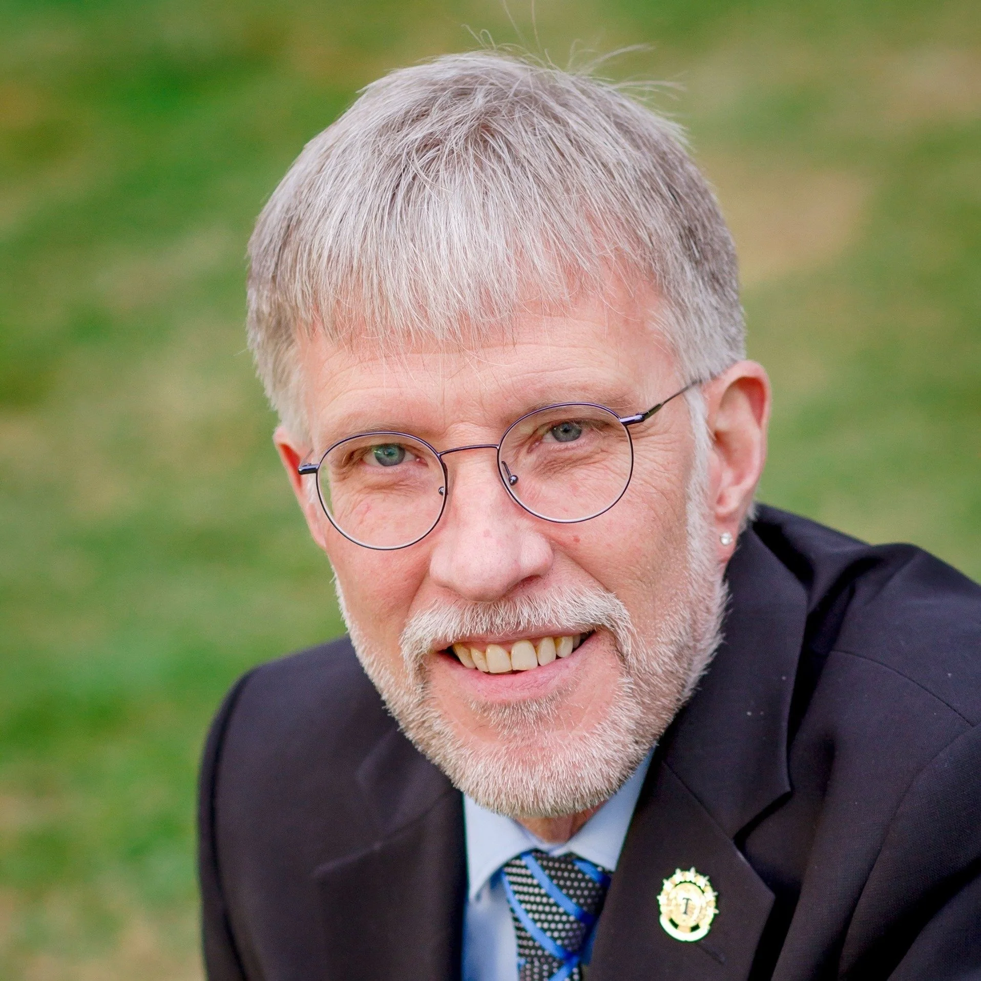 Close-up of an older man with grey hair and glasses, smiling, dressed in a suit and tie, outdoors with a blurred green background.