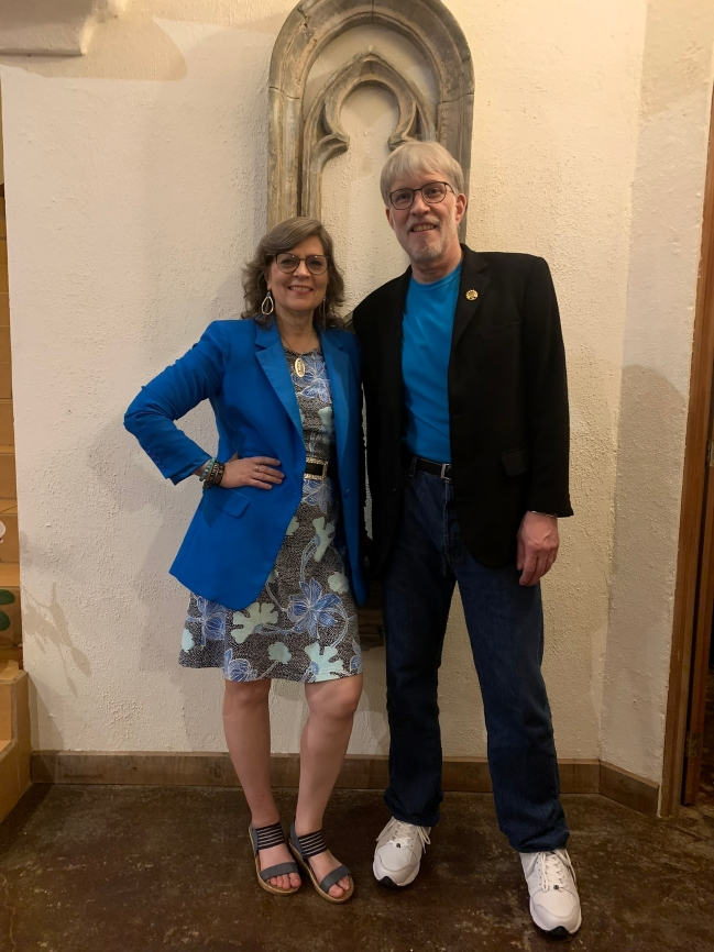 A woman and a man standing together indoors, smiling for the photo. The woman is wearing a blue blazer, a patterned dress, and sandals, while the man is dressed in a black blazer, blue shirt, jeans, and sneakers. They are standing in front of a wall with a decorative arched niche.