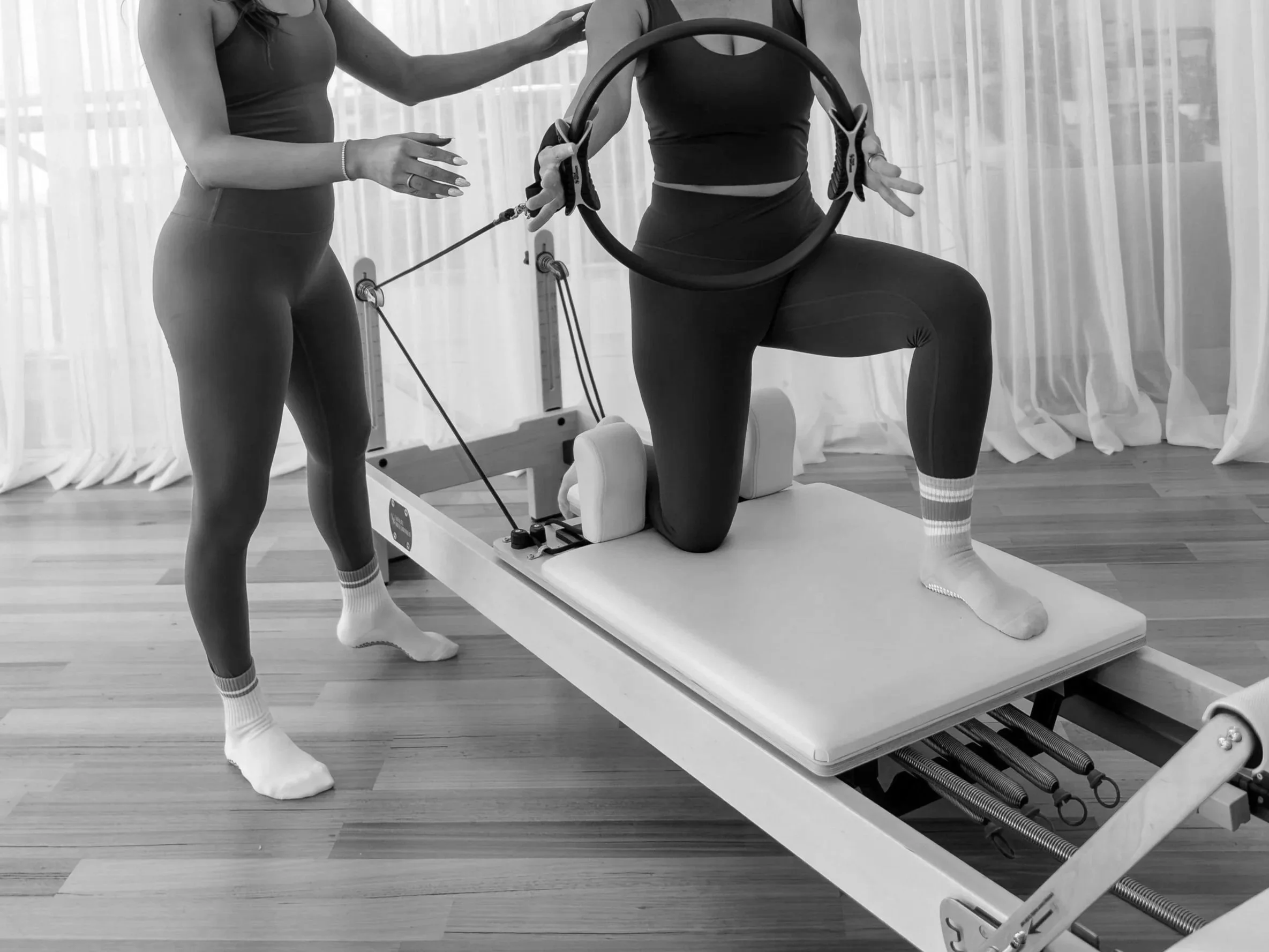 Two women in athletic clothing are practicing pilates in a fitness studio with large windows and curtains. One woman is kneeling on the reformer machine while the other is standing beside her, gently touching her shoulder and giving instructions.