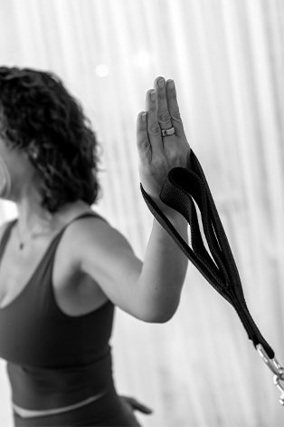 A woman raising her hand with a black strap or leash attached, likely in a yoga or fitness studio, with curtains in the background.