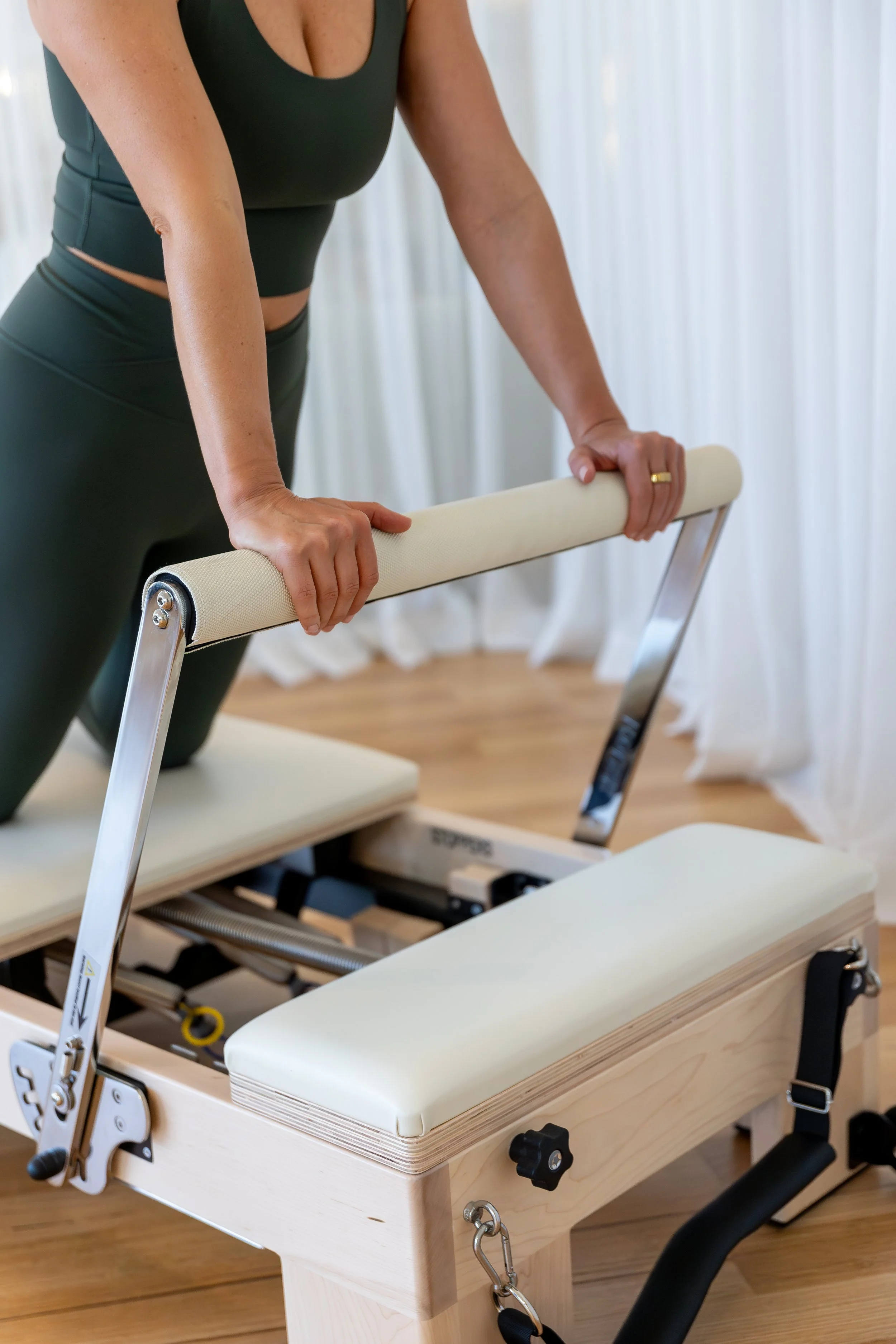 A person is doing exercise on a Pilates reformer with a padded headrest, footbar, and adjustable springs, in a room with wooden floors and white curtains.