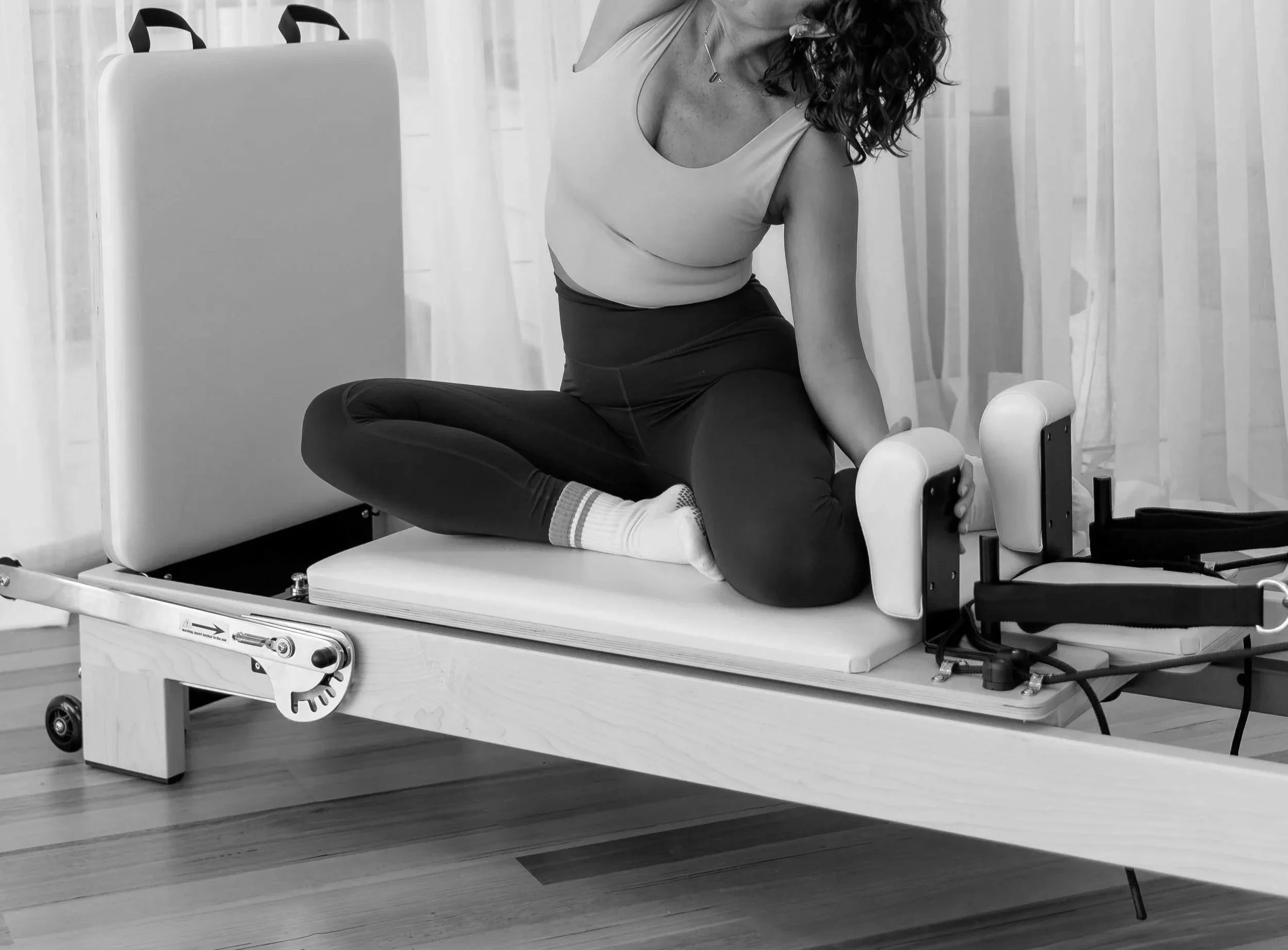 A woman with curly hair seated on a medical examination table in a clinical setting, performing a physical therapy or rehabilitation exercise with a device that supports her leg.