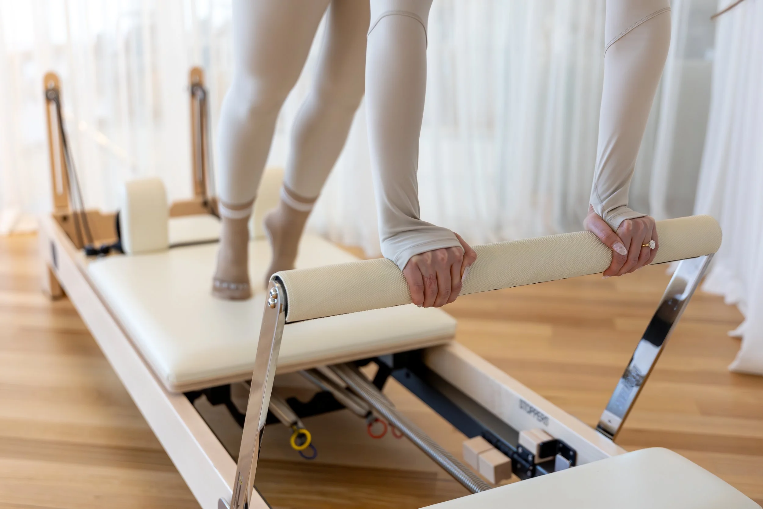 A person in beige leggings is on a Pilates reformer machine, holding a padded bar with both hands while performing a exercise. The reformer has a padded platform and adjustable springs.