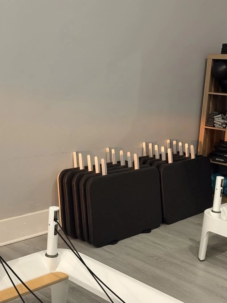 Folded black exercise mats leaning against a wall, with a small shelf containing black weights and rolled-up mats to the right.