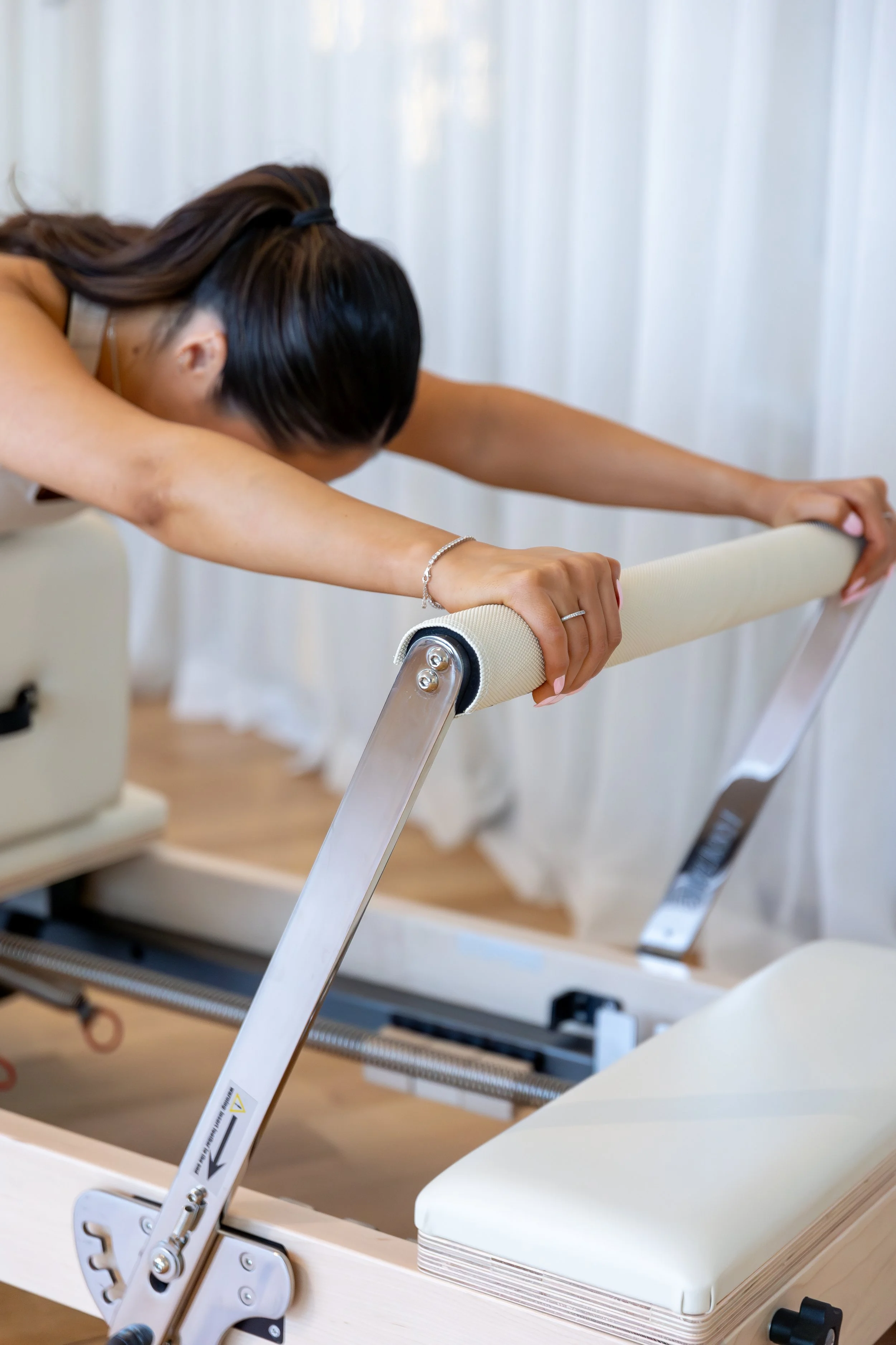 A person performing a stretch on a Pilates reformer machine, with white curtains in the background.