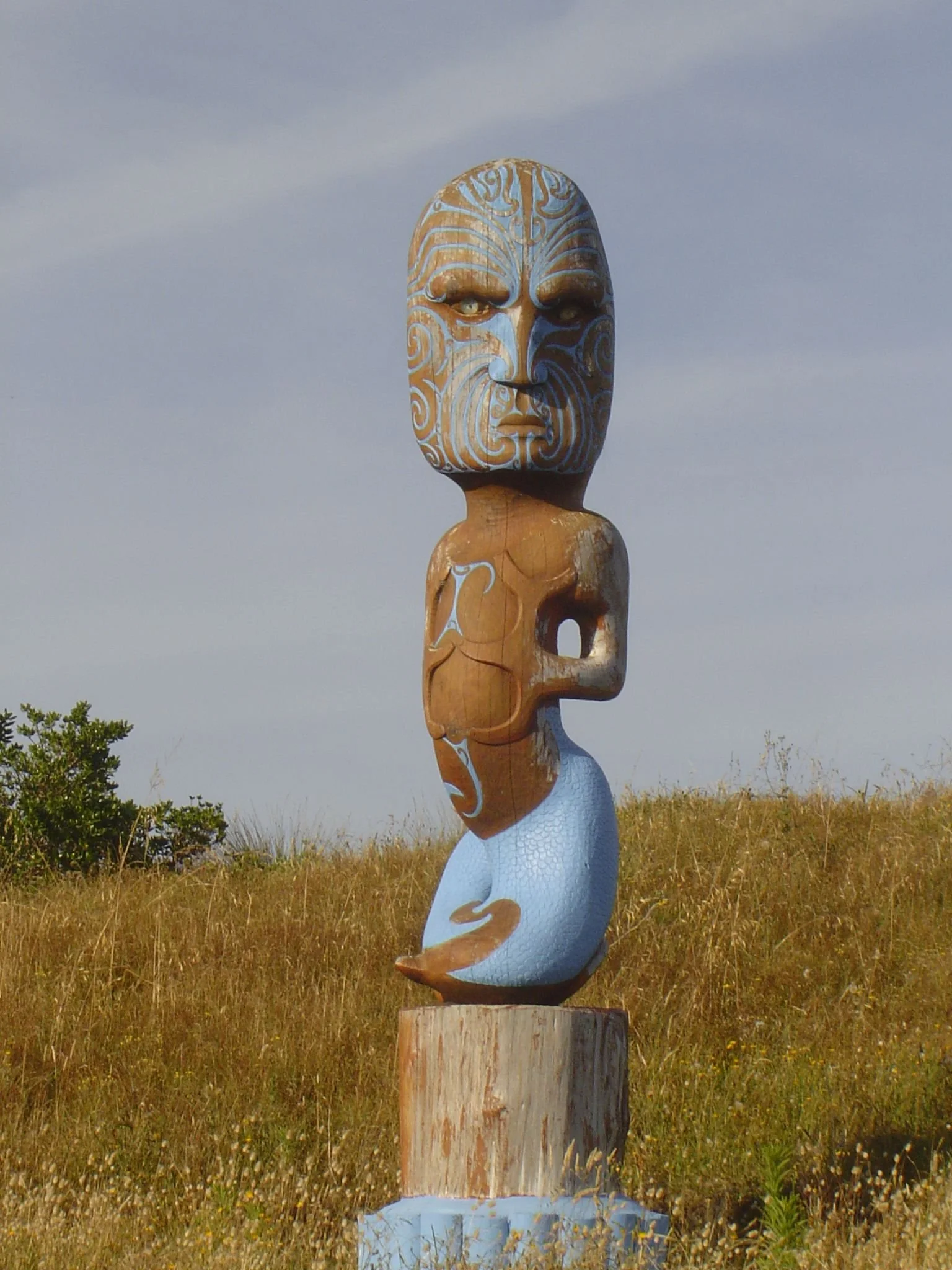 Painted Māori wooden carving with intricate facial tā moko patterns standing in a grassy field