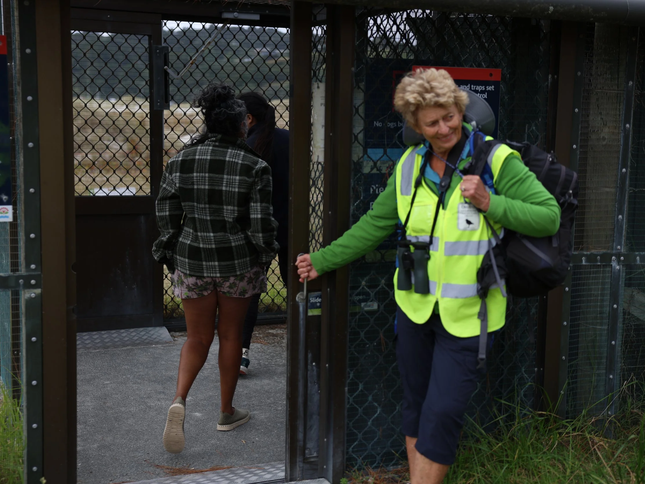 Wildlife sanctuary worker in a high-visibility vest holds open a secured gate as visitors enter an animal enclosure.