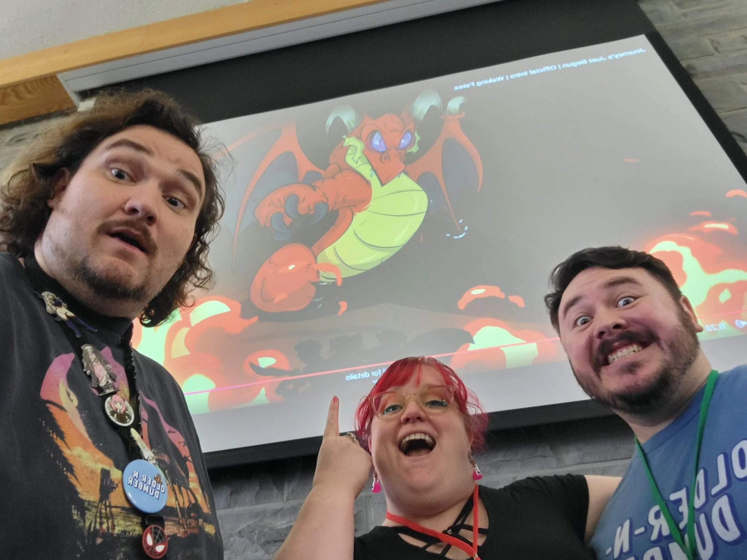 Group of four people taking a selfie in front of a screen displaying a cartoon dragon with orange, green, and blue colors