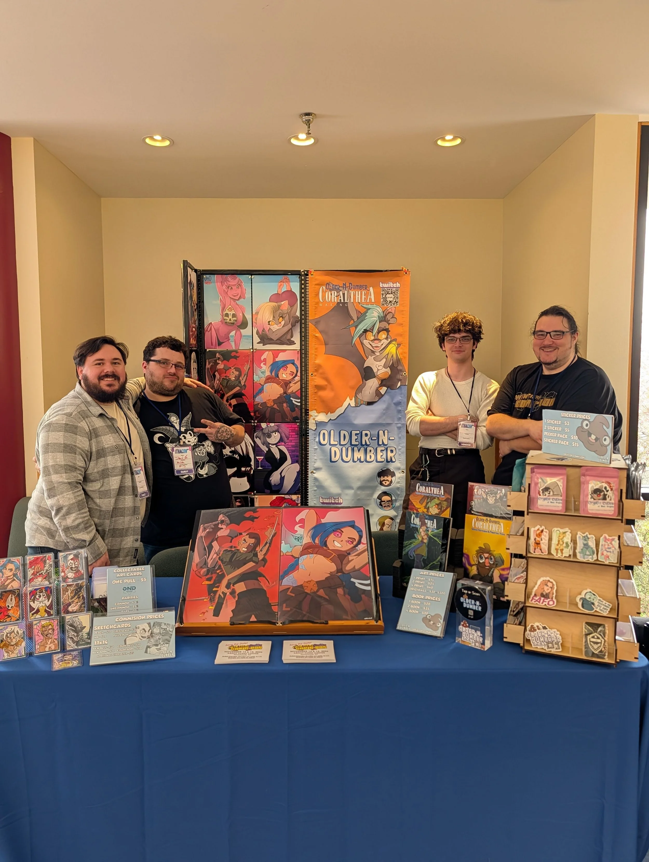 A group of four men standing behind a table at a convention or event. The table displays various artwork, prints, and stickers related to a fantasy or cartoon style game called 'Older-N Dumber'. Behind them are colorful banners featuring animated characters, with one banner reading 'CoralThea' and another 'Older-N Dumber'. The men are smiling and looking at the camera, wearing casual clothing and name badges.