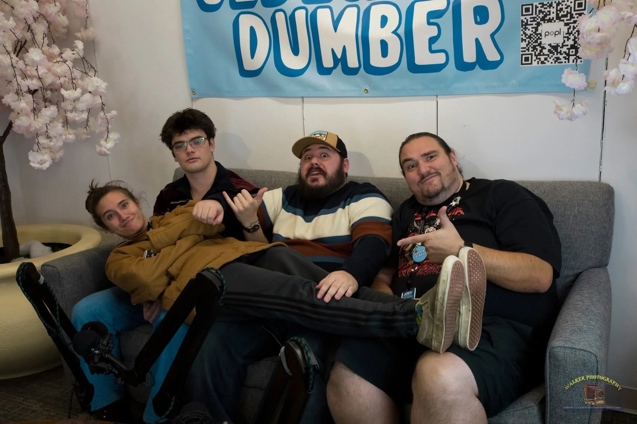 Four people sitting on a couch in front of a sign that says 'OlderNDumber' with a QR code. Two trees with pink flowers are on either side of the sign.