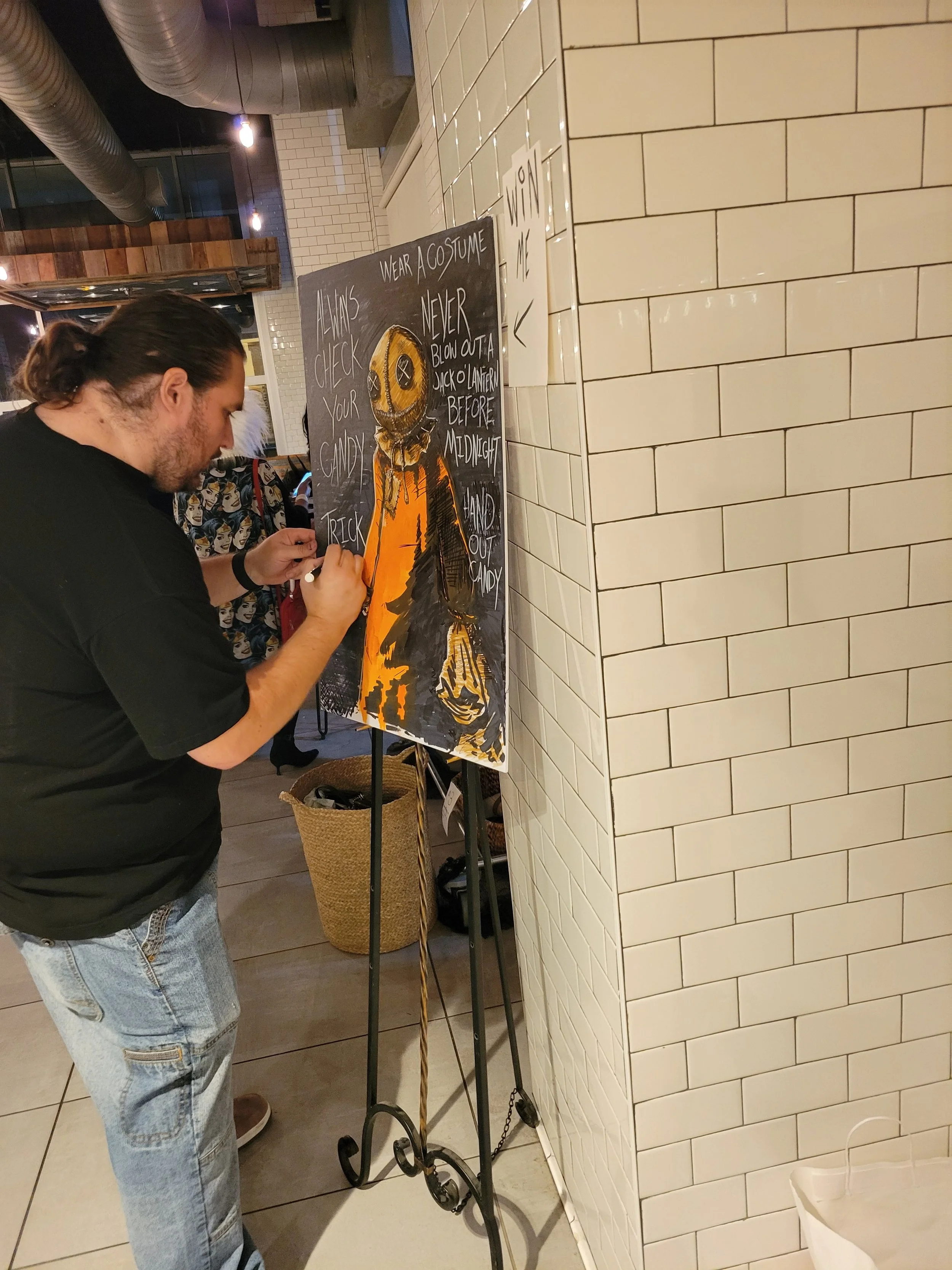 A man drawing or writing on a Halloween-themed chalkboard sign in a restaurant or cafe. The sign has a pumpkin illustration and instructions for Halloween safety, including checking candy and not blowing out jack-o'-lanterns before midnight.