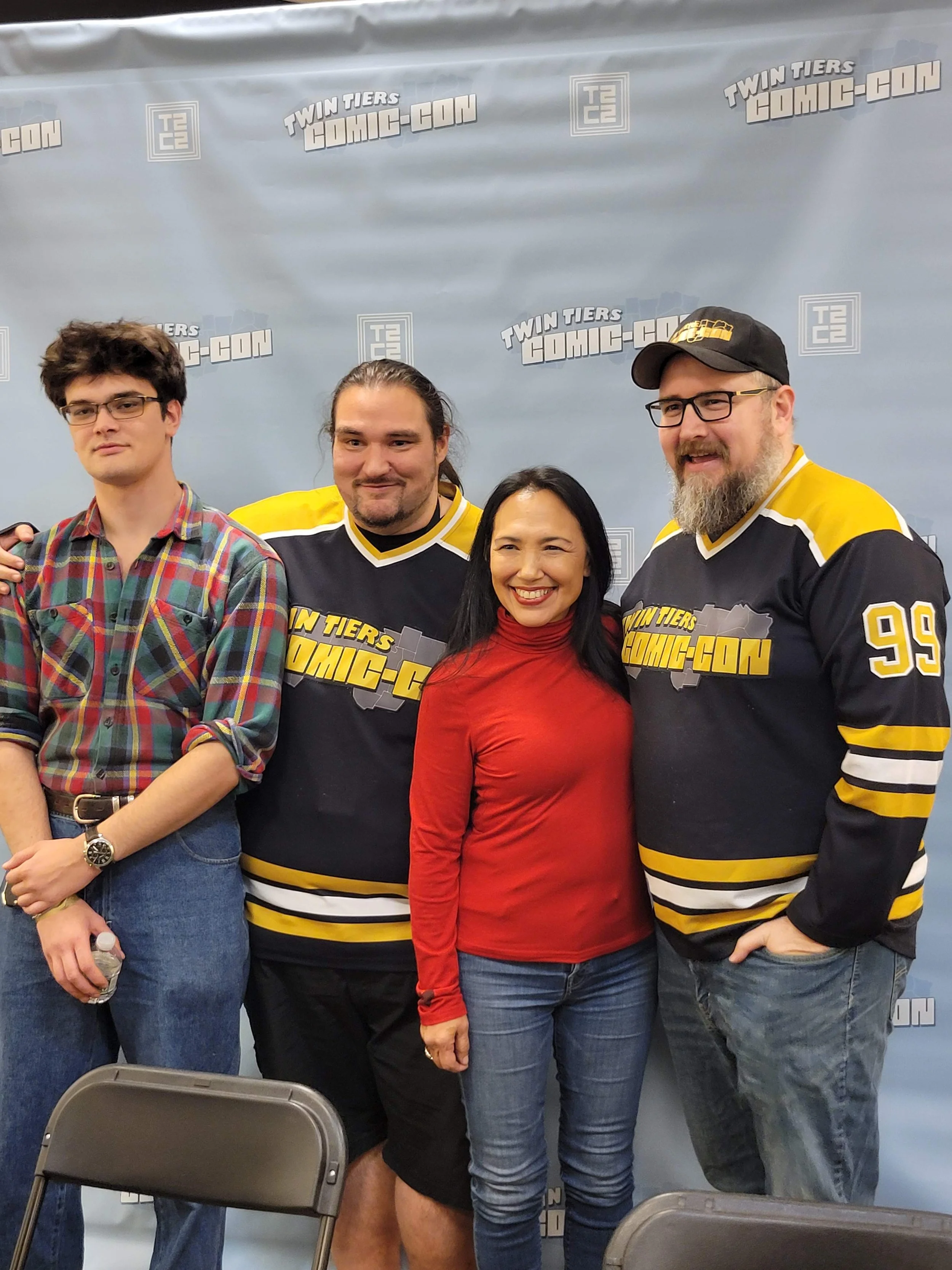 Four people, three men and one woman, standing together at a Comic-Con event. The three men are wearing black and yellow hockey jerseys with the words 'Twin Tiers Comic-Con' on them. The woman is wearing a red top. They are posing in front of a backdrop with the Twin Tiers Comic-Con logo.