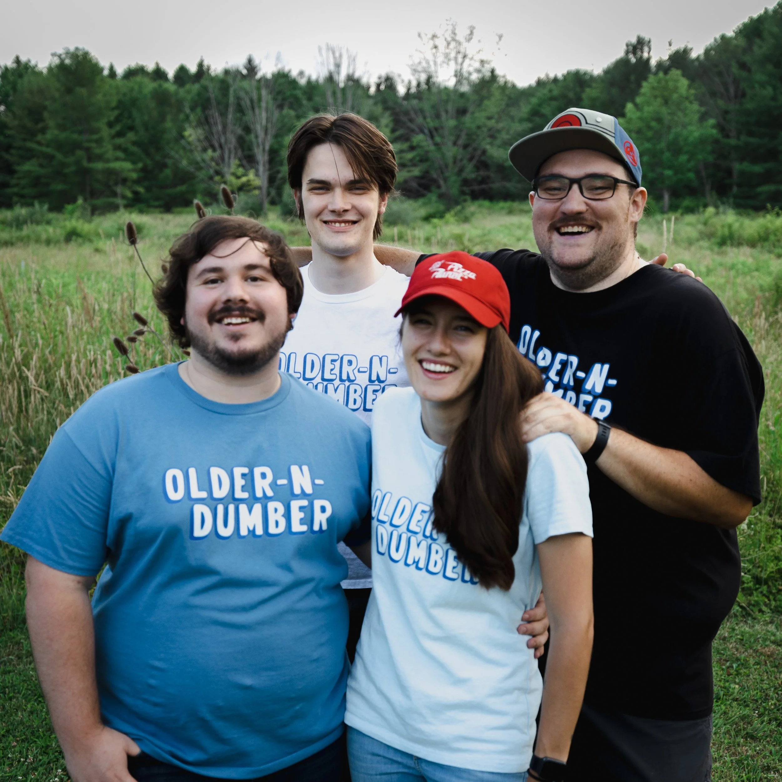 Group of four friends standing outdoors in a grassy field with trees in the background, smiling and taking a selfie. Some are wearing sunglasses and casual clothes, with several wearing blue and white shirts that say 'OLDERNDUMBER'.