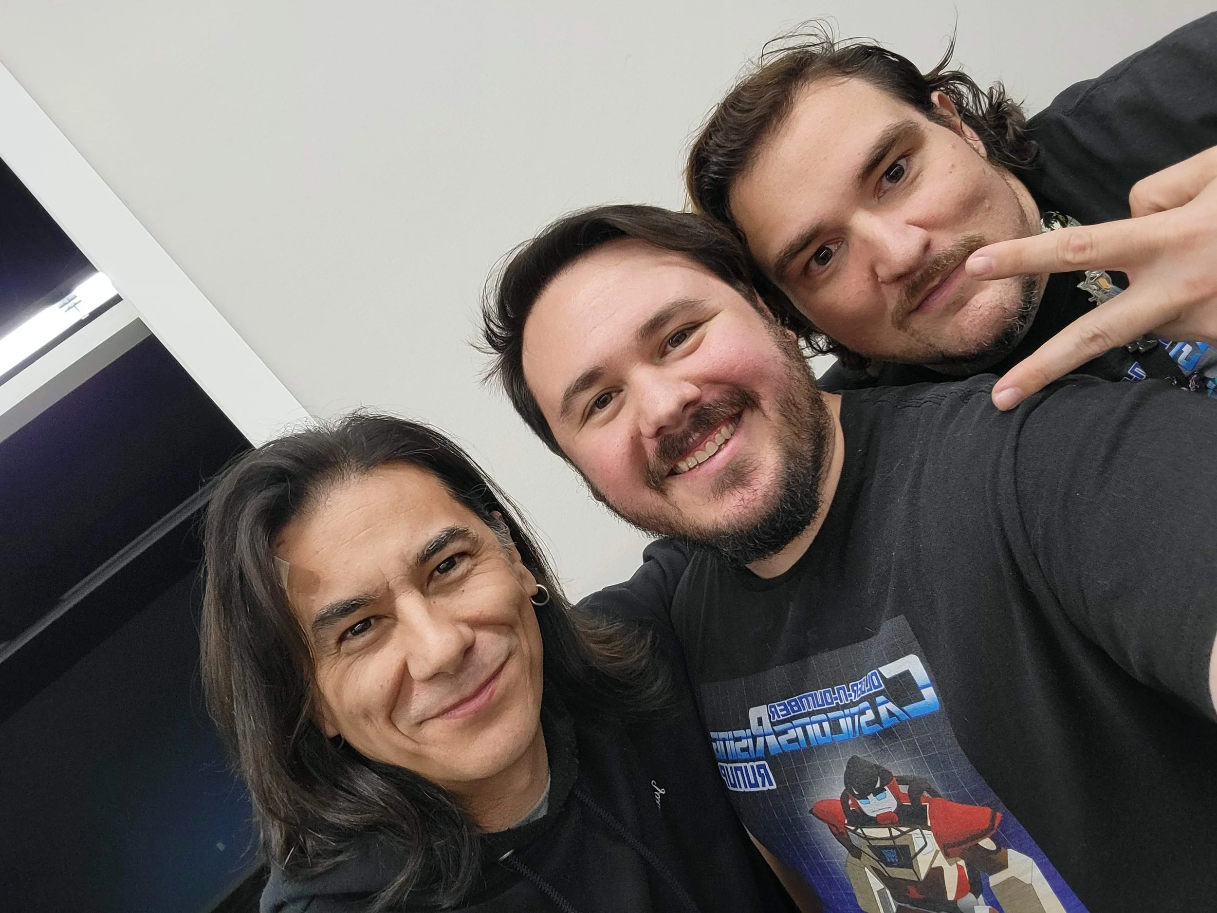 Three people taking a selfie in an indoor setting, smiling and making peace signs, with a plain white wall in the background.