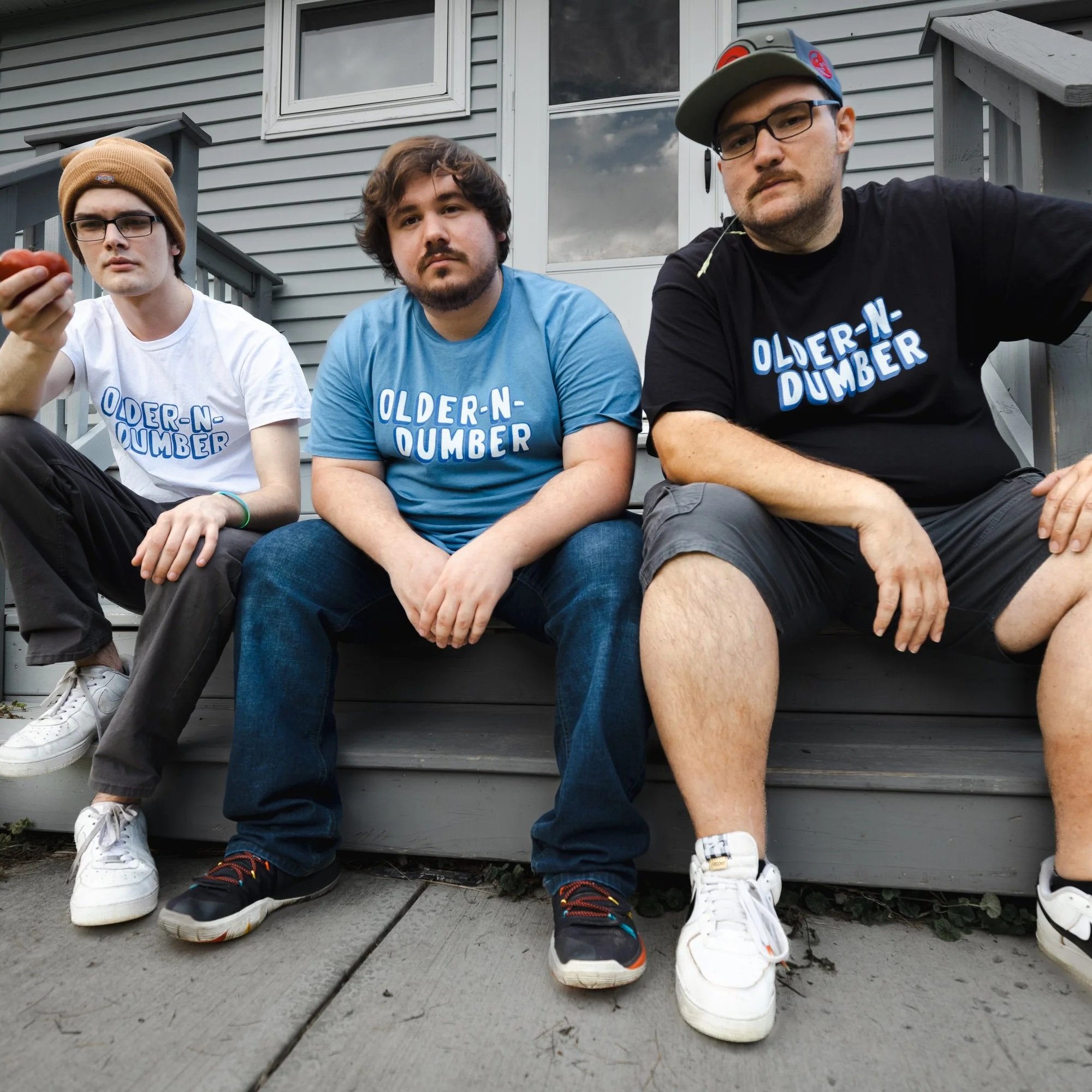 Three young men sitting on a porch step in front of a house with gray siding. They wear casual clothing, including t-shirts that say 'OLDER-N-DUMBER'. The young man on the right wears a caps and glasses, the one in the middle has wavy brown hair, and the one on the left wears a brown beanie and glasses. All are sitting with their legs crossed or hanging down.