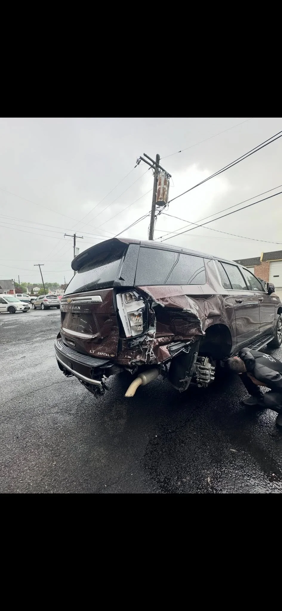 Damaged SUV after crash, missing rear wheel, with a person working nearby, in a parking lot under a cloudy sky.