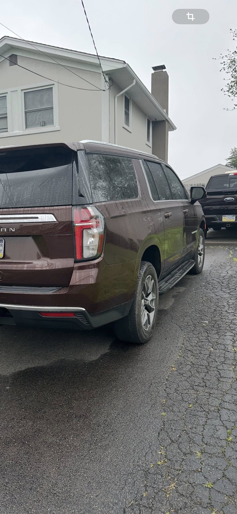 A maroon SUV parked on a cracked asphalt lot in front of a white residential building with multiple windows and a chimney. There are other vehicles parked nearby, and the sky is overcast.