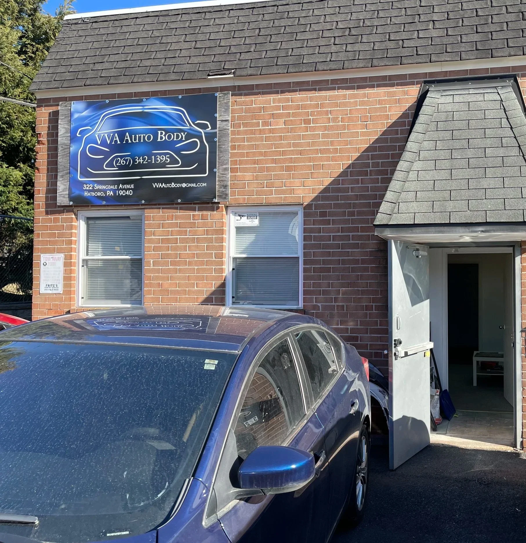 A brick building with a sign for VVA Auto Body, including a car shape logo and contact information. Car parked in front, open garage door visible.