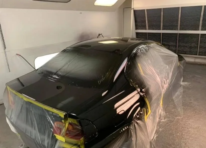 Black car covered in plastic sheeting and tape inside a garage, likely undergoing a paint job or protection.