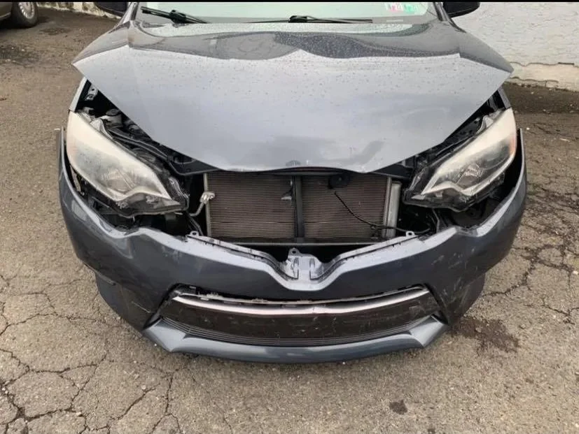 Front view of a damaged gray car with a missing front grille and exposed radiator, on a cracked asphalt surface.