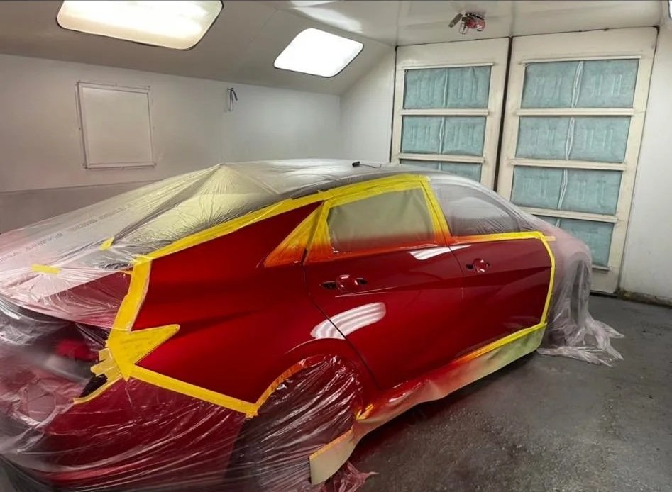 Red car being painted inside a garage with plastic sheeting and yellow masking tape.
