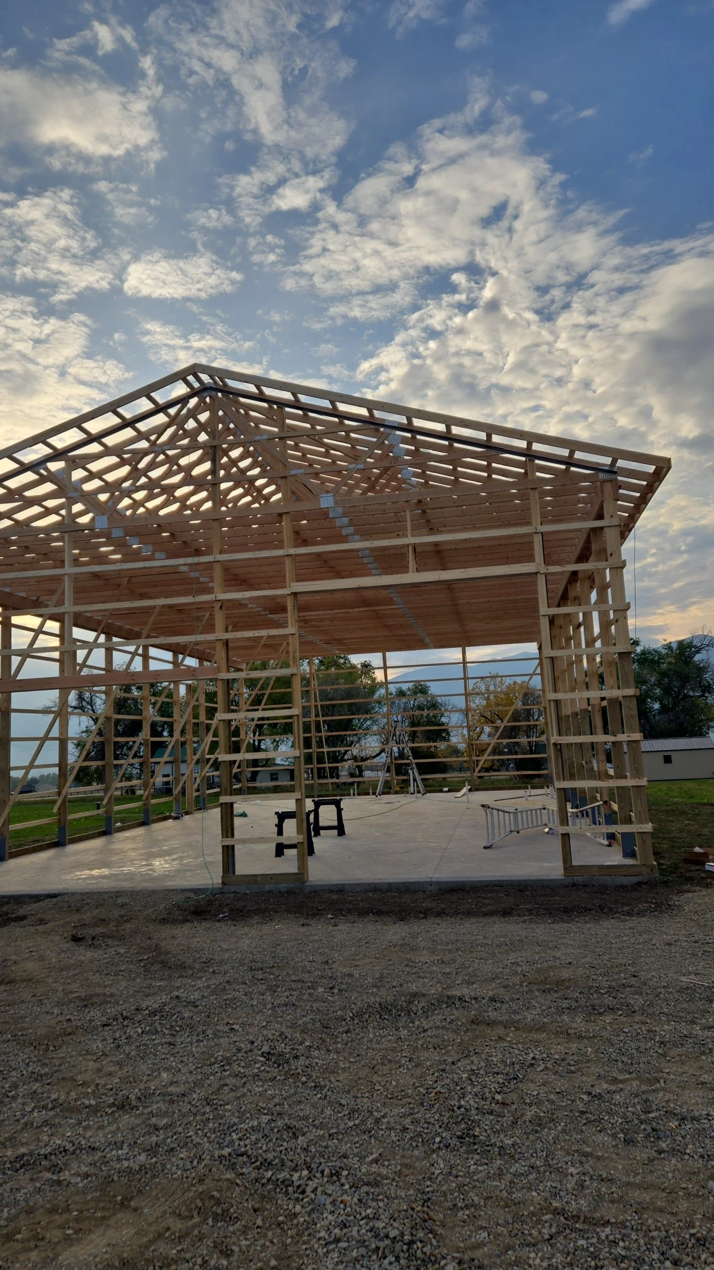 Wooden house frame under construction with a high ceiling, supported by vertical and horizontal beams, set against a partly cloudy sky in the background.