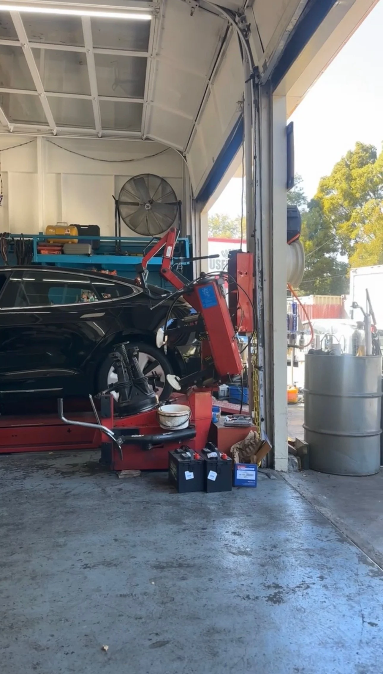 Inside an auto repair garage with a black car on a lift, various tools, equipment, and a large industrial fan visible. Parts and supplies are organized around the workspace.