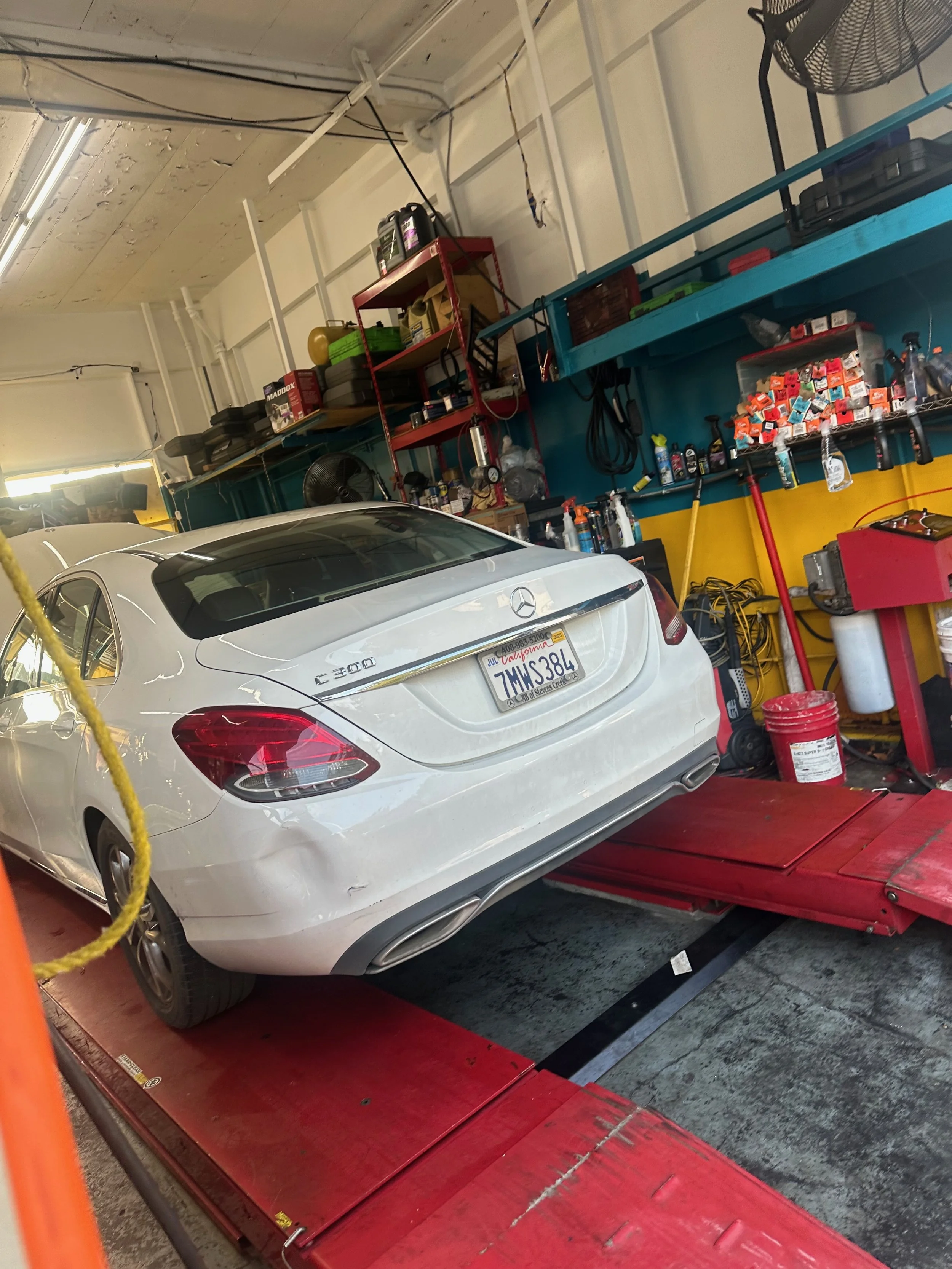 A white Mercedes-Benz E300 sedan on a red car lift in a garage or auto repair shop, with shelves of tools and supplies on the wall behind it.