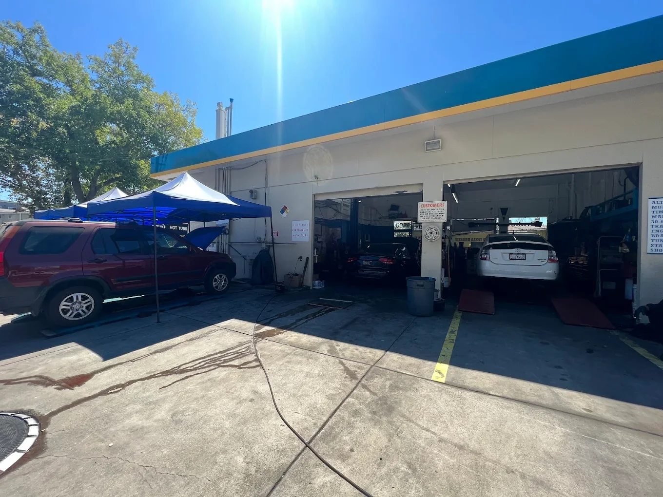 Auto repair shop with several cars on lifts and parked outside, sunny day with blue sky and green trees