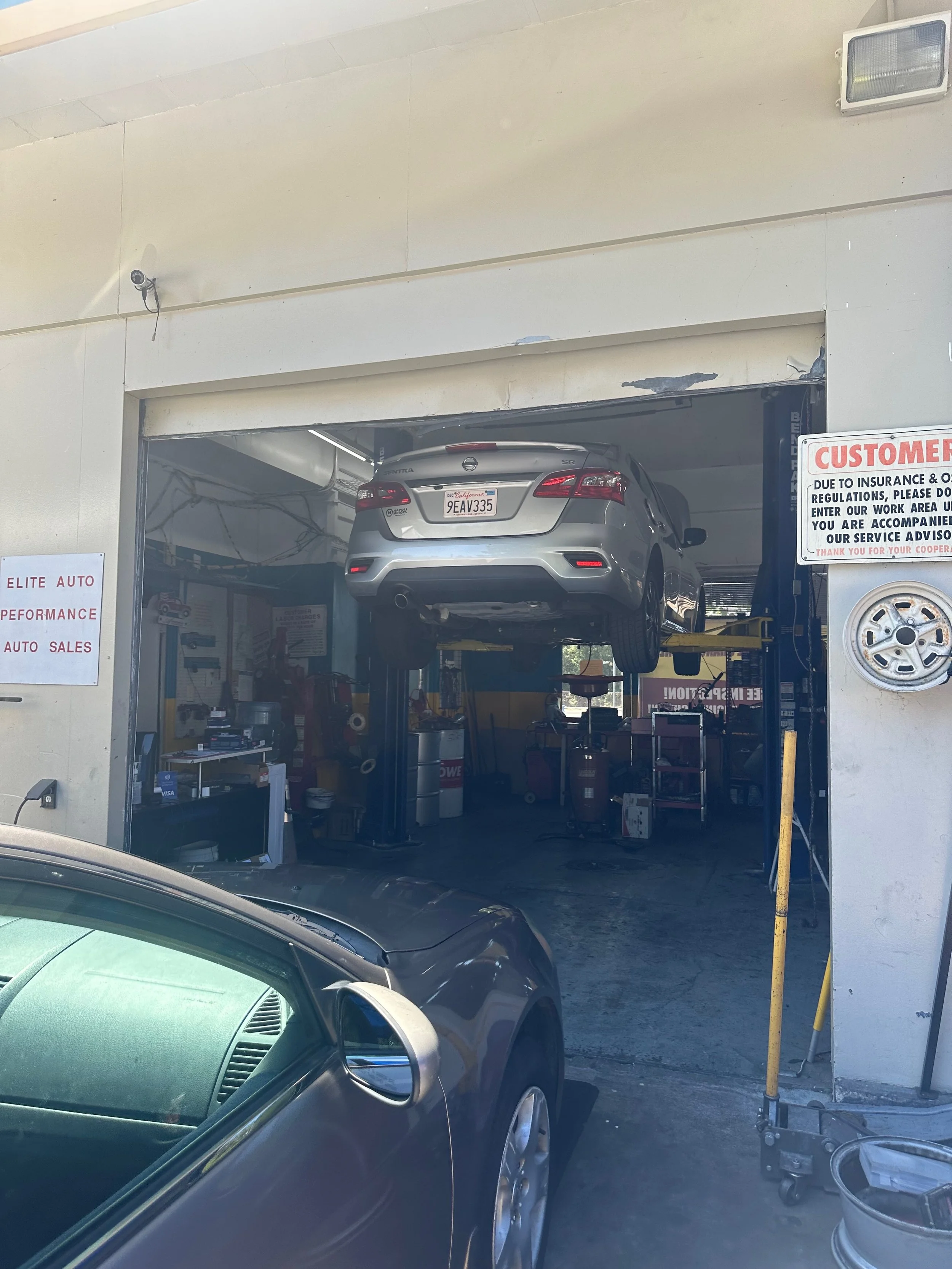 A car lifted on an auto mechanic lift inside an automotive repair shop. The workshop has signs and equipment visible around.