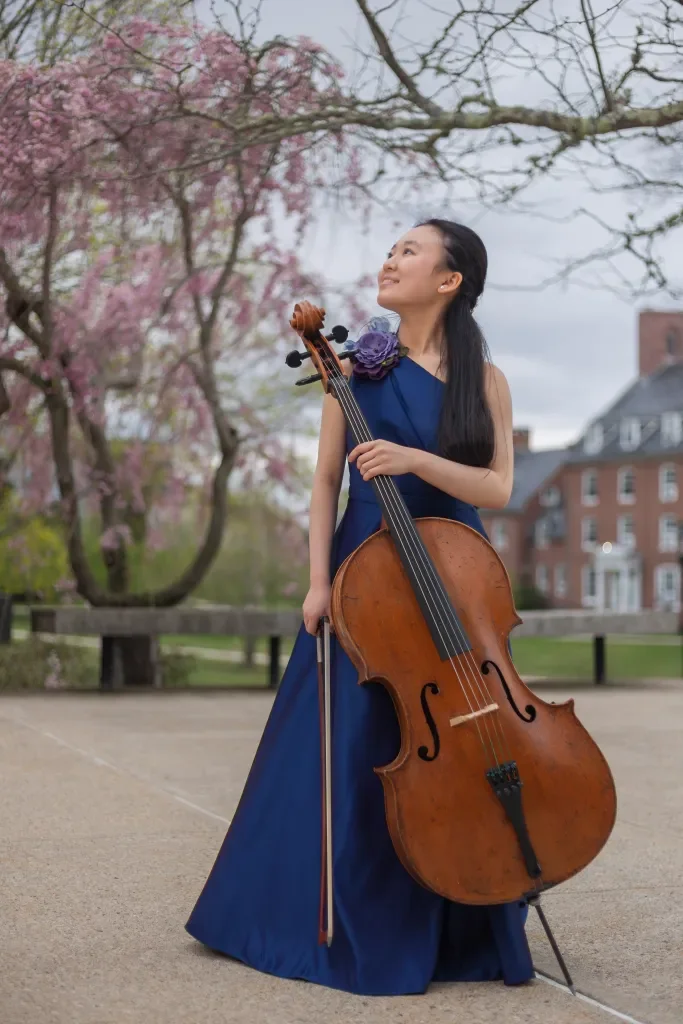 A woman in a blue evening gown holding a cello and bow, standing outdoors near blooming pink trees.