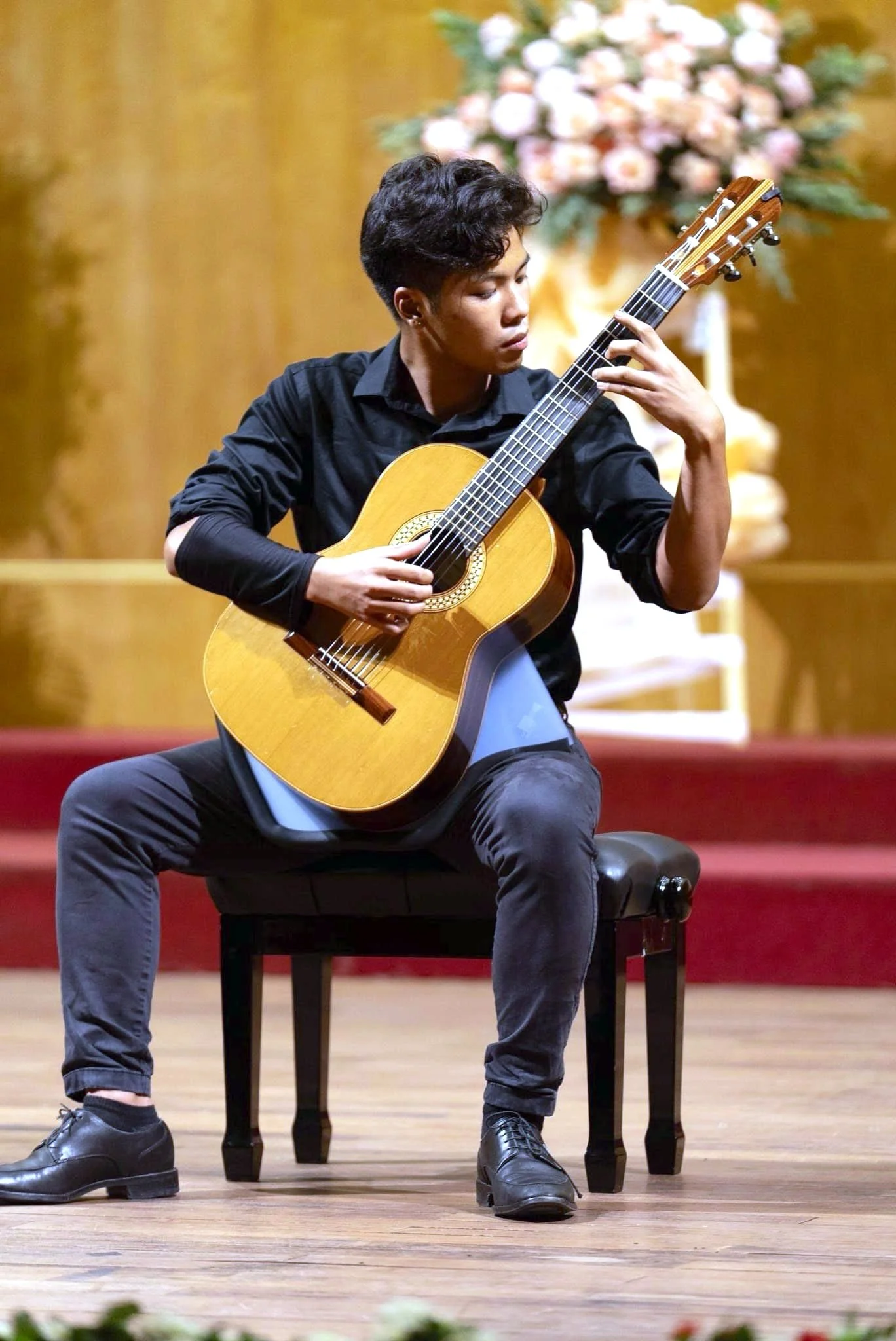 A young person sitting on a chair playing an acoustic guitar on stage with a floral arrangement and wooden panel background.