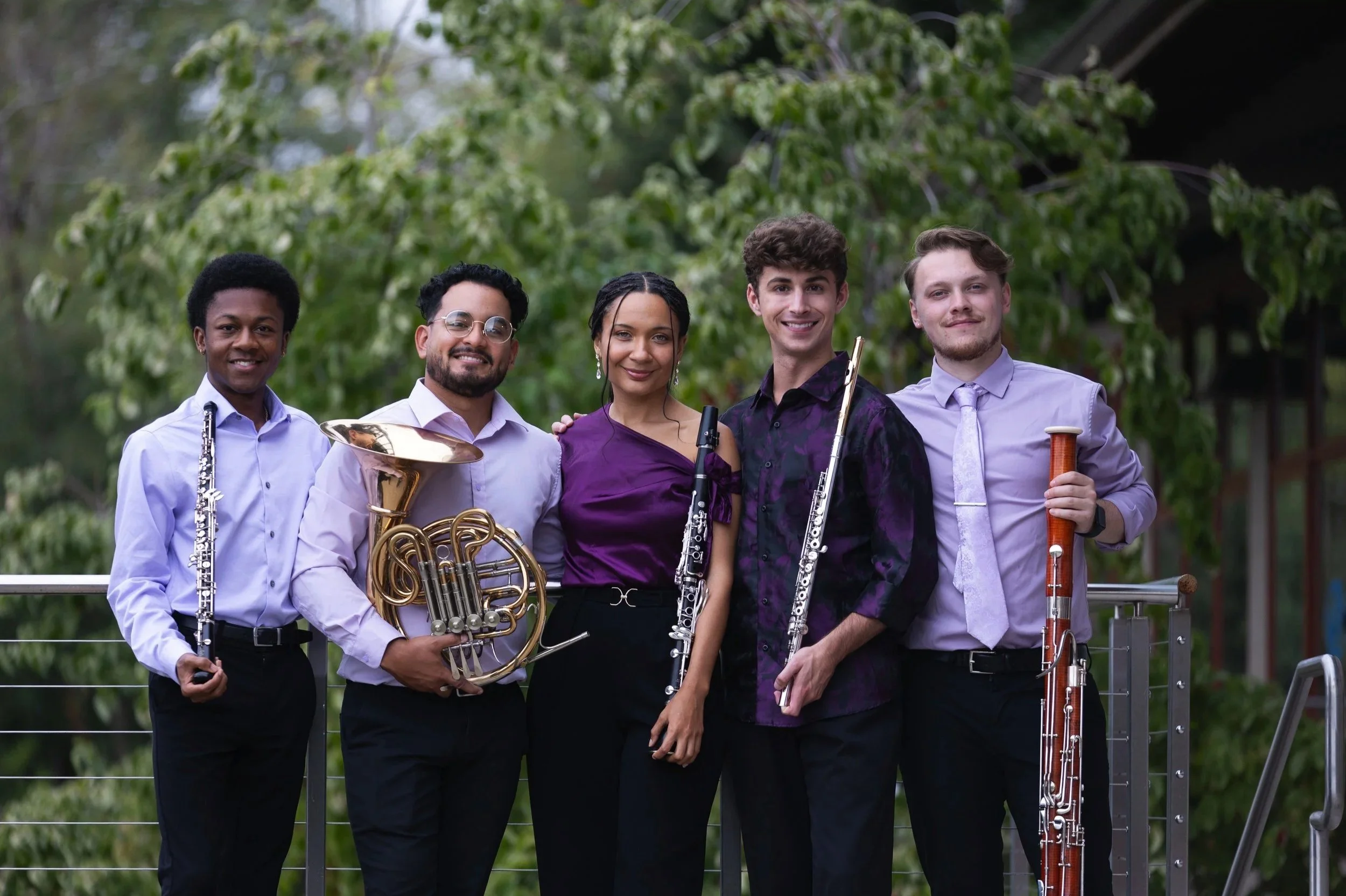 A group of five musicians standing outdoors, holding various musical instruments, smiling at the camera, with green trees and a building in the background.