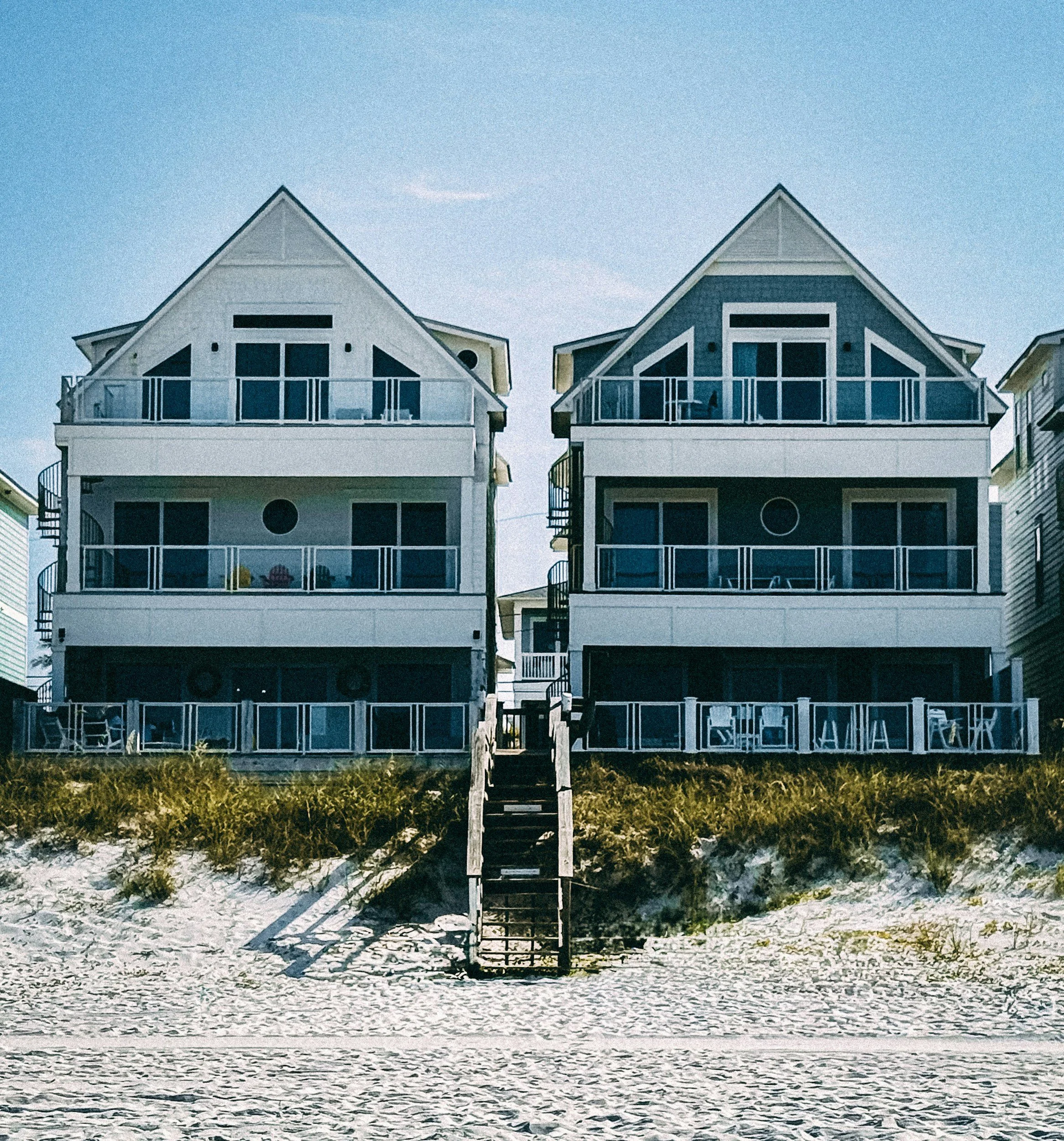 Two multi-story beach houses with balconies and a central staircase leading to the sandy shore.