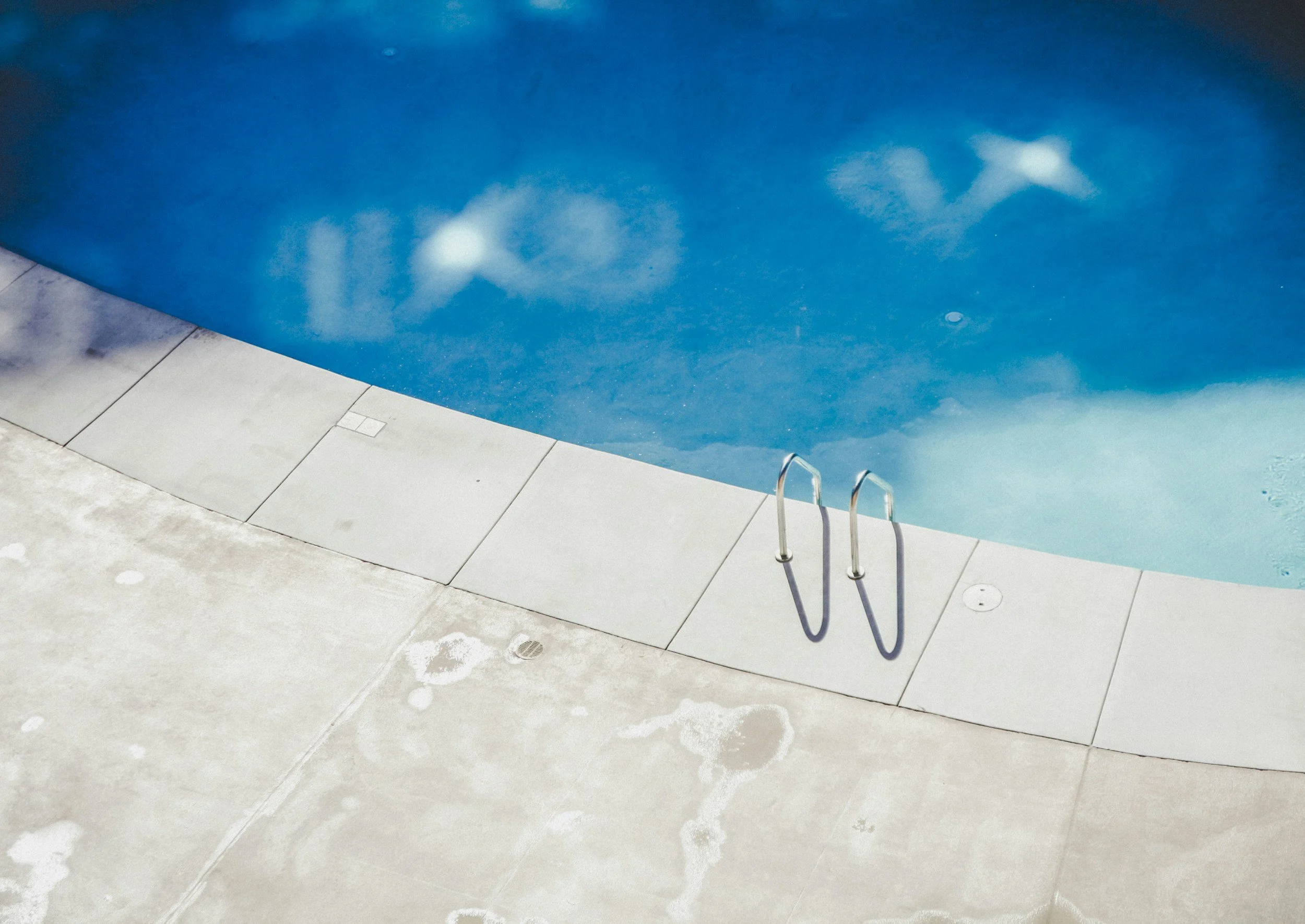 Empty swimming pool with metal handrails, surrounded by light-colored tiled area, and some poolside drainage covers.
