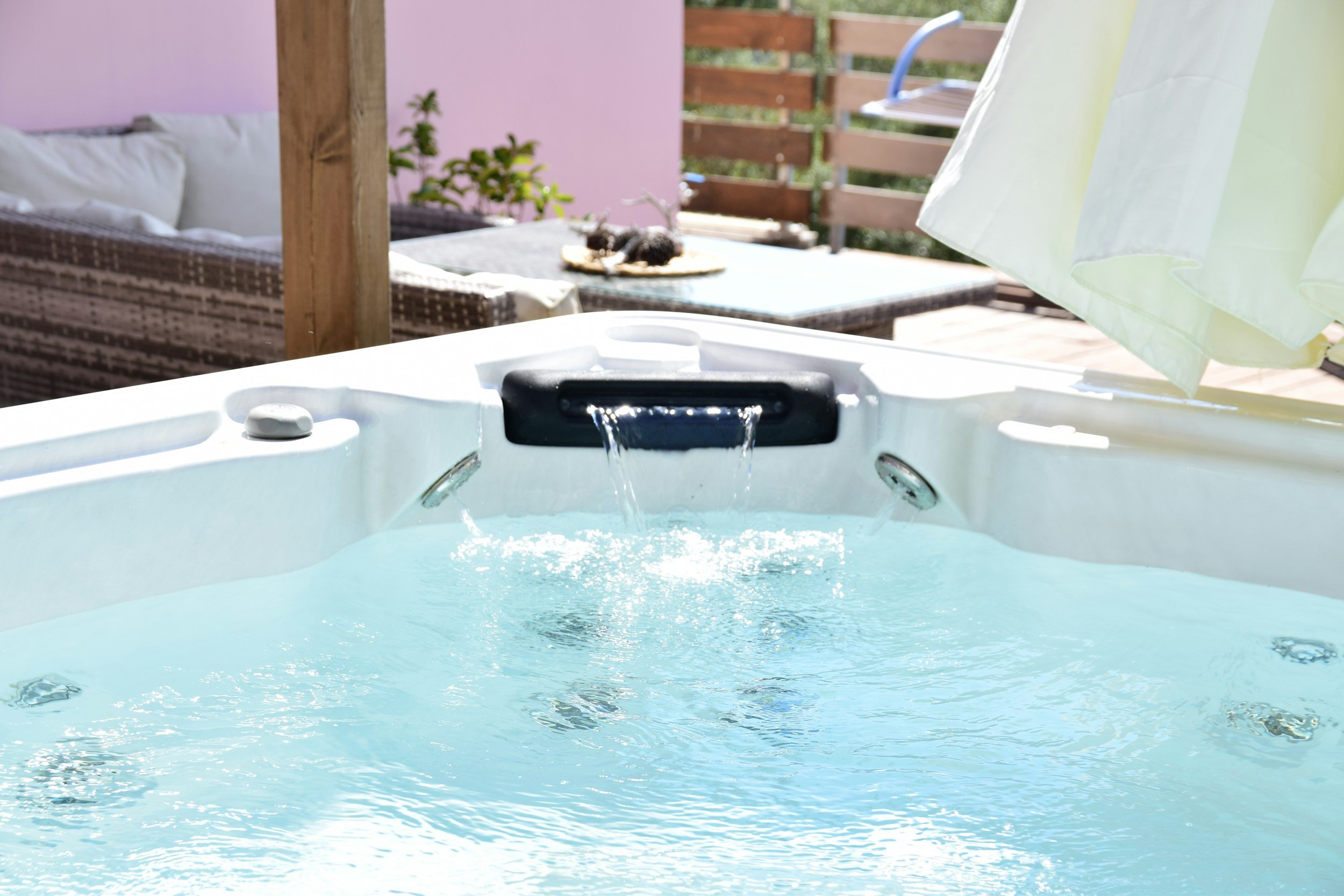 View of an outdoor hot tub filled with water, with a waterfall feature, on a sunny day. In the background, there is a wooden fence, a pink wall, a table with a small potted plant, and a black and white cat lying on a cloth on the table. Part of a green and white umbrella is visible on the right side.