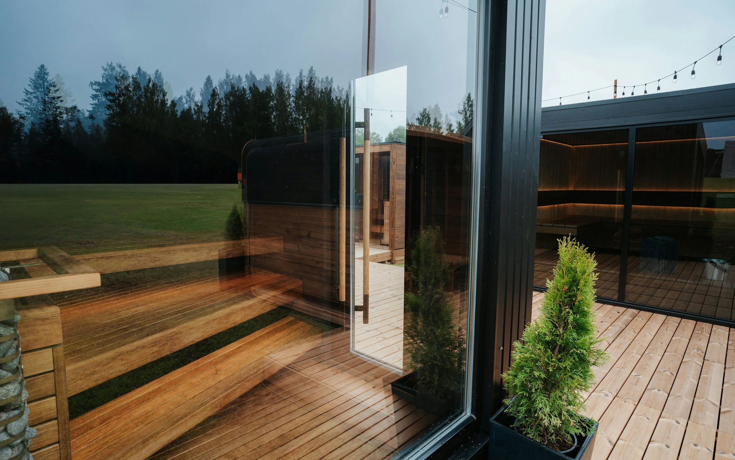 View through a glass door showing an outdoor wooden deck with potted plants, string lights, green grass, and trees in the background.