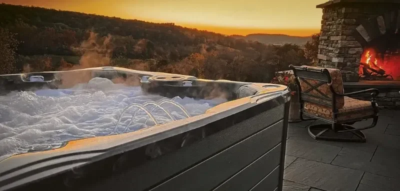 Hot tub outdoors at sunset with a fire in a stone fireplace and a cozy chair, overlooking a hilly landscape.