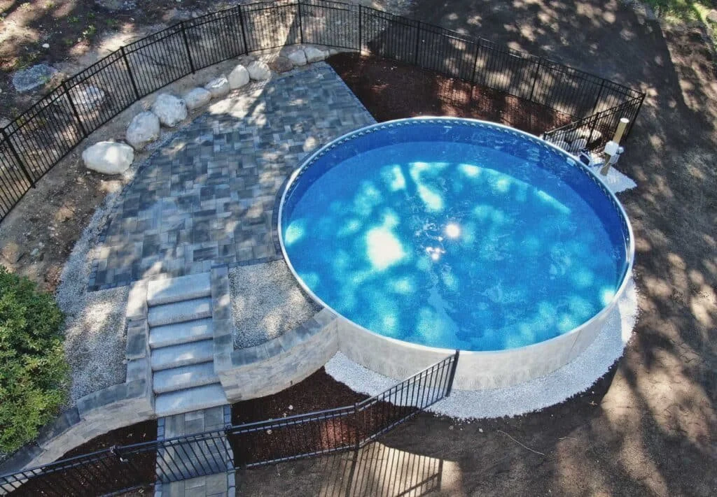 A round above-ground swimming pool filled with blue water, enclosed by a black metal fence, with stone steps leading up to it and a small paved area nearby surrounded by trees and rocks.